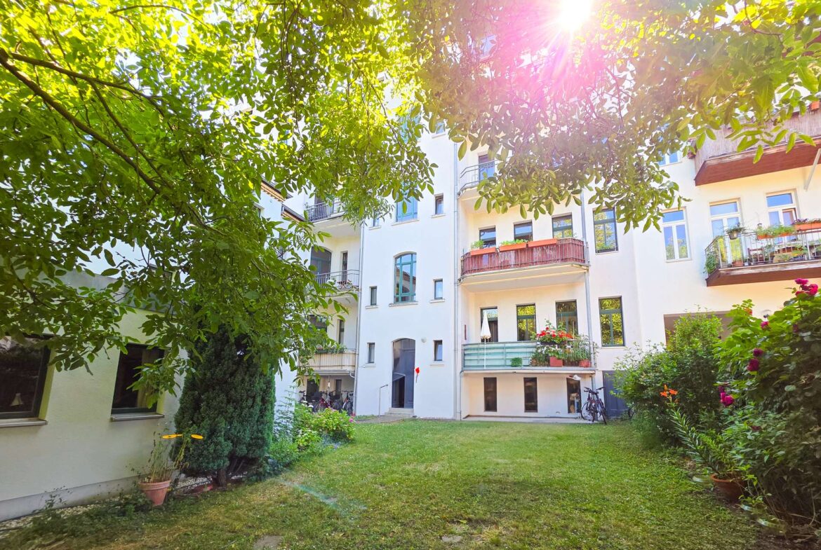 Ein sonniger Innenhof mit grünem Gras, Bäumen und Pflanzen, umrahmt von einem weißen Wohnhaus mit Balkonen und großen Fenstern. Das Sonnenlicht fällt durch die Blätter und schafft eine helle, einladende Atmosphäre.