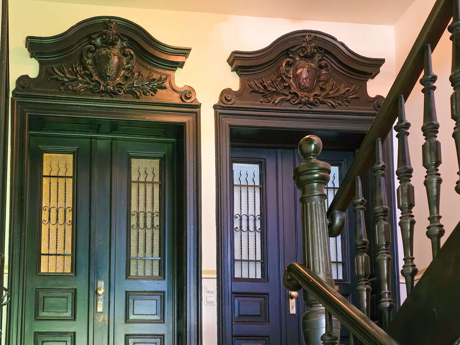 Zwei verschnörkelte Türen aus dunklem Holz, über denen kunstvolle Schnitzereien mit schildartigen Motiven angebracht sind. Rechts neben den Türen führt eine Holztreppe mit gedrechselten Balustern hinauf. Die Beleuchtung ist warm und einladend.