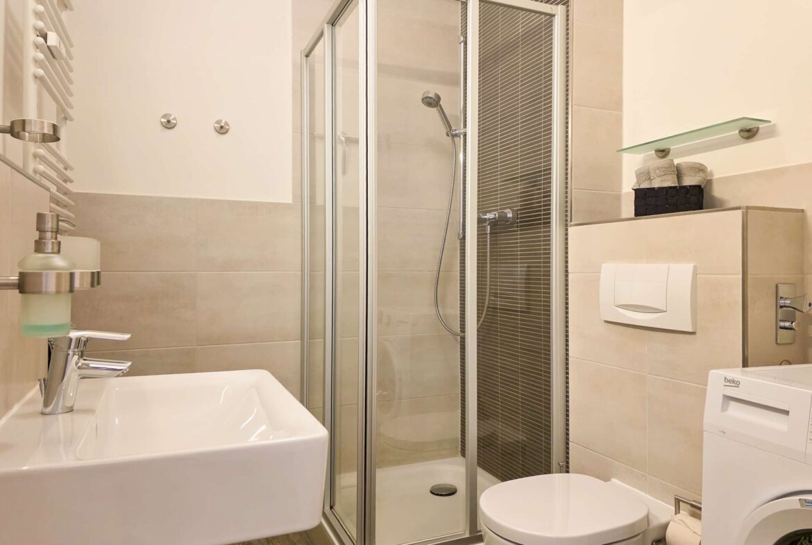 Modernes Badezimmer mit Glasduschabtrennung, Wandwaschbecken, Toilette, Waschmaschine und Regalen mit gefalteten Handtüchern und Toilettenartikeln vor beigen Fliesen.
