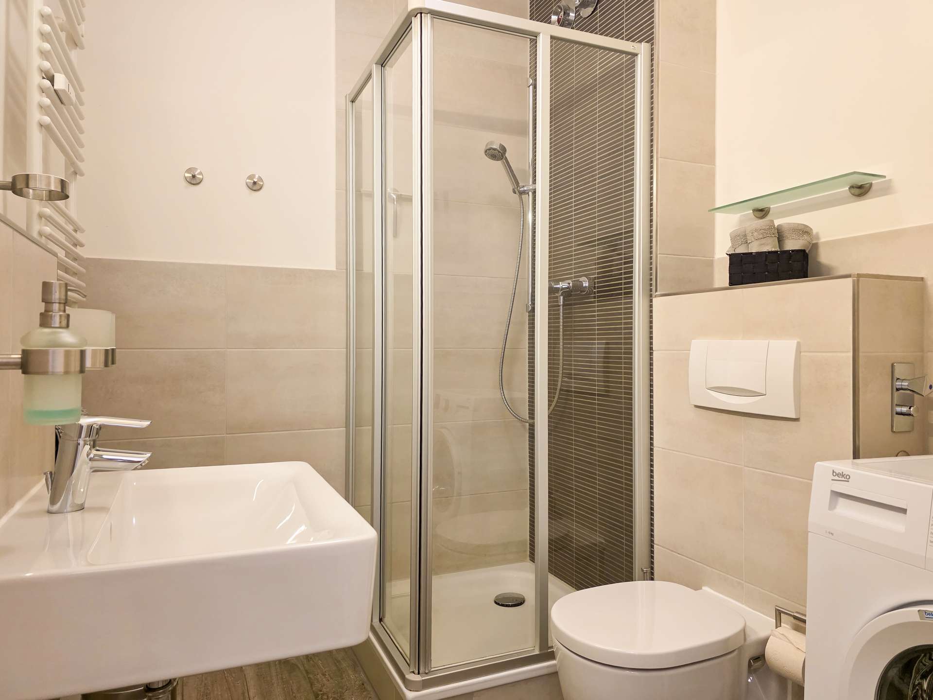 Modernes Badezimmer mit Glasduschabtrennung, Wandwaschbecken, Toilette, Waschmaschine und Regalen mit gefalteten Handtüchern und Toilettenartikeln vor beigen Fliesen.