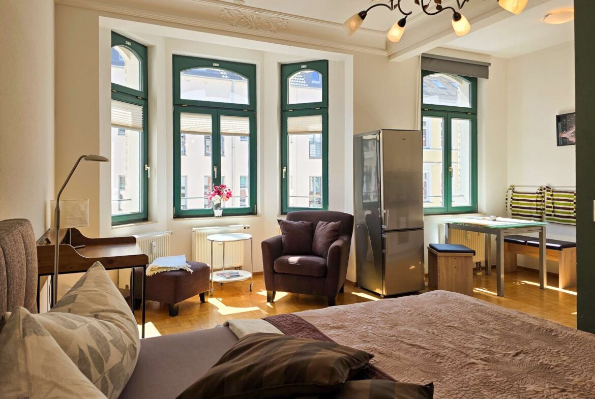Helles, gemütliches Schlafzimmer mit großen, grün gestrichenen Fenstern, einem Sessel mit Fußstütze, einem kleinen Tisch, einem Schreibtisch, einer Küchenzeile und einem Bett mit Kissen und einer braunen Bettdecke. Sonnenlicht strömt durch die Fenster.