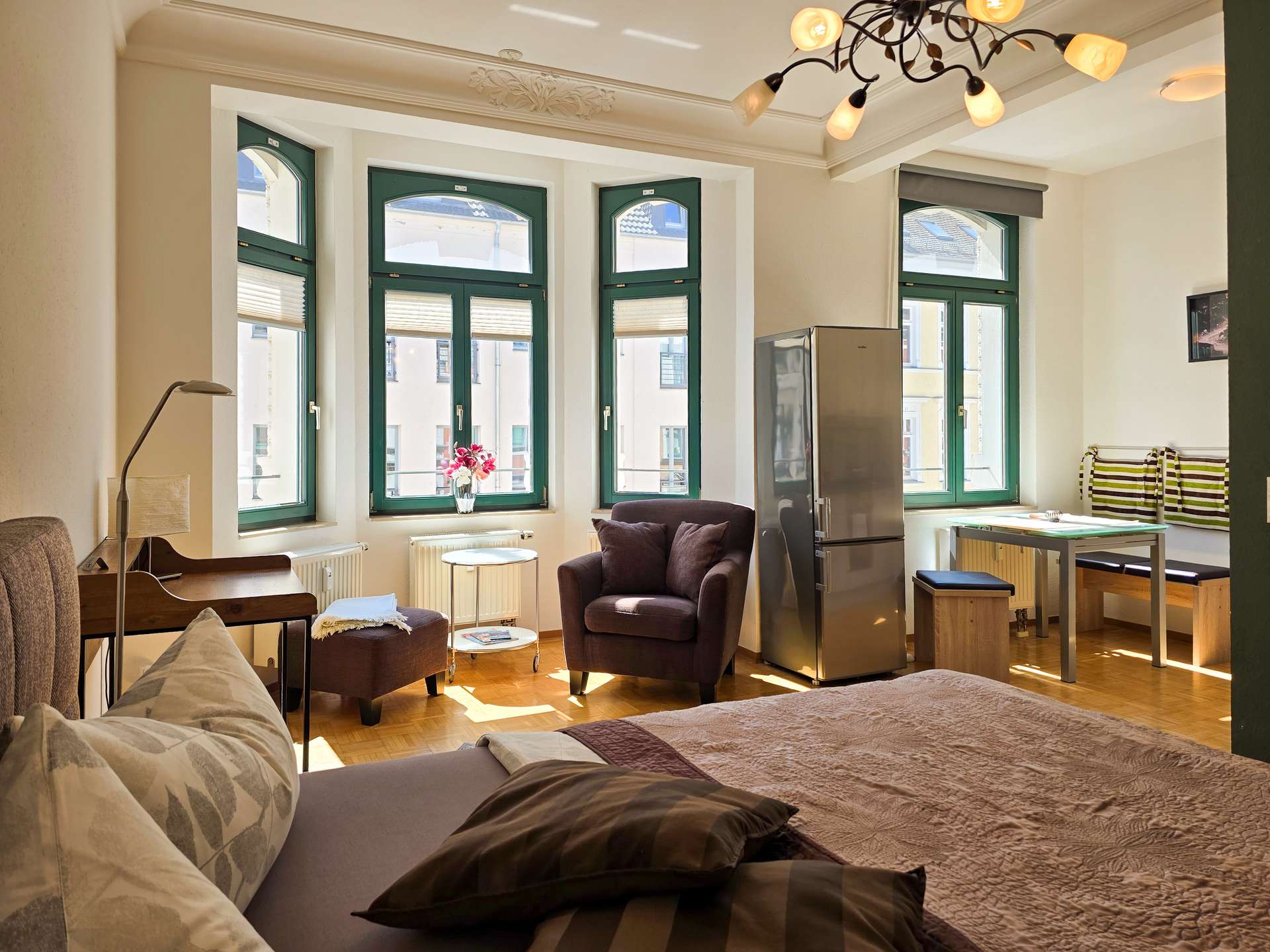 Helles, gemütliches Schlafzimmer mit großen, grün gestrichenen Fenstern, einem Sessel mit Fußstütze, einem kleinen Tisch, einem Schreibtisch, einer Küchenzeile und einem Bett mit Kissen und einer braunen Bettdecke. Sonnenlicht strömt durch die Fenster.