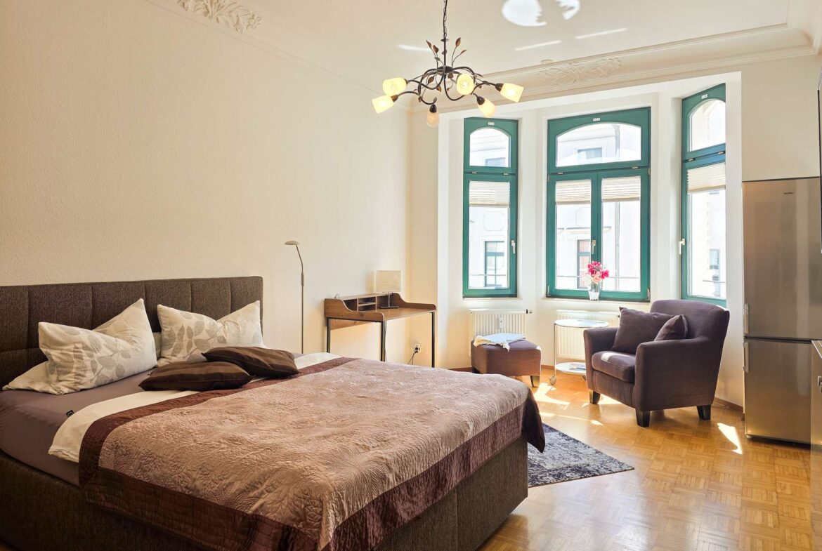 Helles Schlafzimmer mit großem Bett, Kissen und brauner Decke, einem gemütlichen Sessel und einem kleinen Tisch am Fenster, grün gestrichenen Fenstern, Kronleuchter, Schreibtisch, Lampe und einem Kühlschrank in der Ecke auf Holzfußboden.