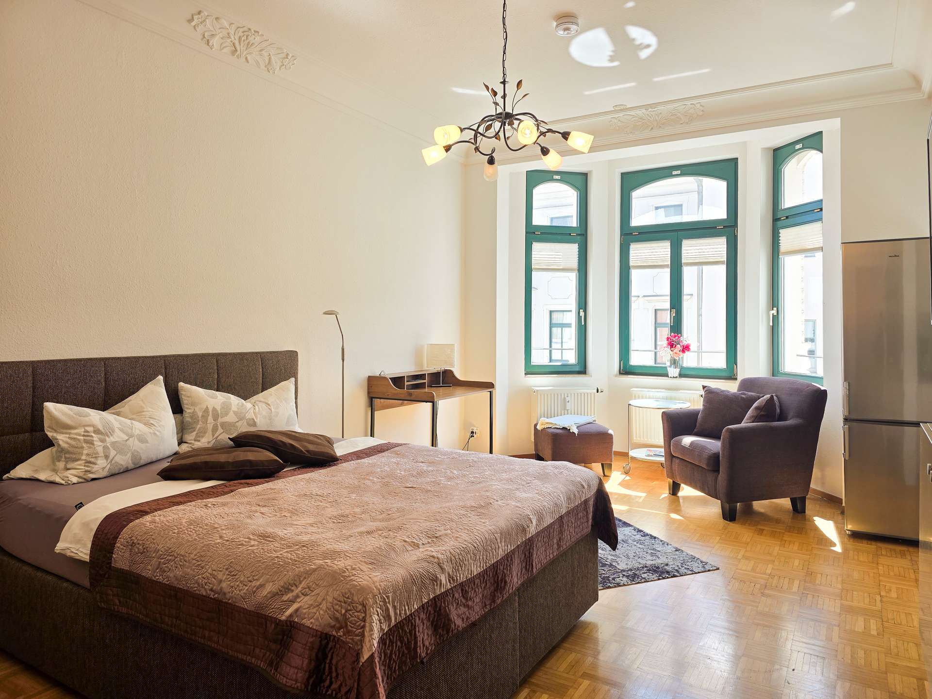 Helles Schlafzimmer mit großem Bett, Kissen und brauner Decke, einem gemütlichen Sessel und einem kleinen Tisch am Fenster, grün gestrichenen Fenstern, Kronleuchter, Schreibtisch, Lampe und einem Kühlschrank in der Ecke auf Holzfußboden.