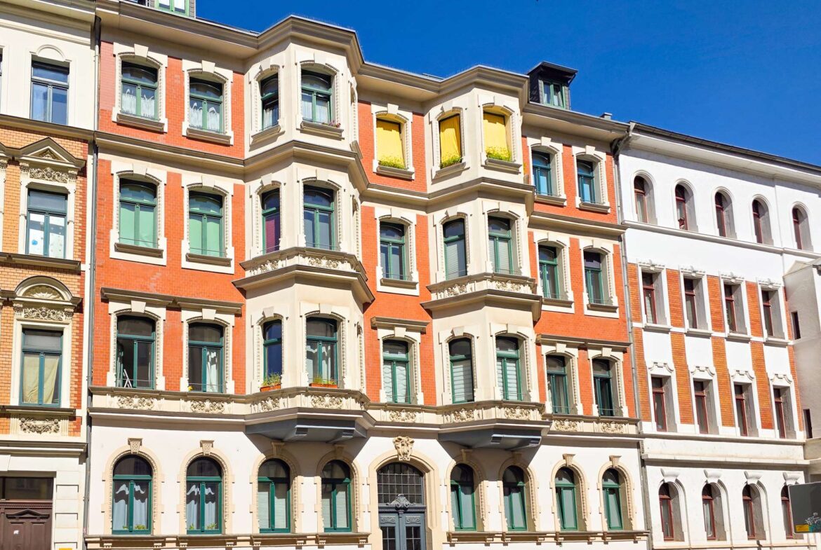 Ein historisches, vierstöckiges Gebäude mit rotem Backstein und cremefarbenen Akzenten, grünen Fensterrahmen, verzierten Balkonen und einer dunklen Eingangstür unter einem strahlend blauen Himmel. In zwei oberen Fenstern sind gelbe Vorhänge zu sehen.