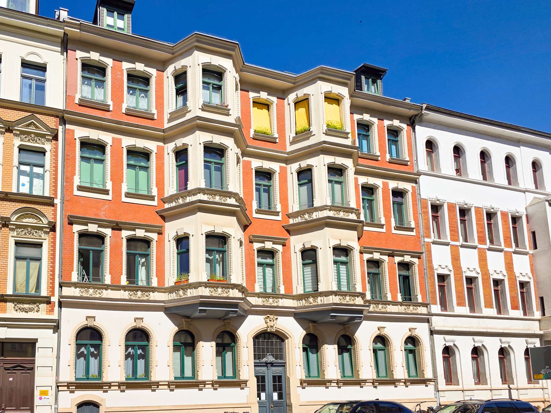 Ein historisches, vierstöckiges Gebäude mit rotem Backstein und cremefarbenen Akzenten, grünen Fensterrahmen, verzierten Balkonen und einer dunklen Eingangstür unter einem strahlend blauen Himmel. In zwei oberen Fenstern sind gelbe Vorhänge zu sehen.