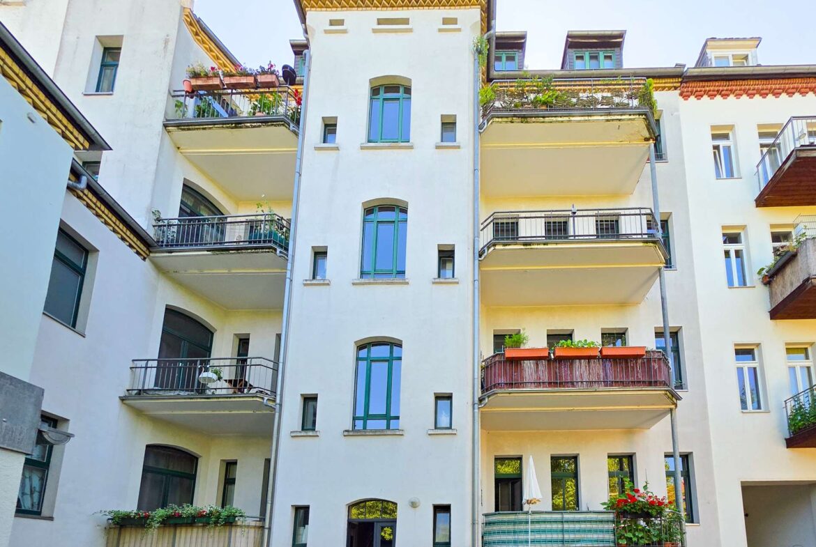 Ein weißes mehrstöckiges Wohnhaus mit Balkonen, die teilweise mit Pflanzen und Blumen geschmückt sind. Die Fassade ist mit großen Fenstern versehen, und auf beiden Seiten befinden sich Nebengebäude.