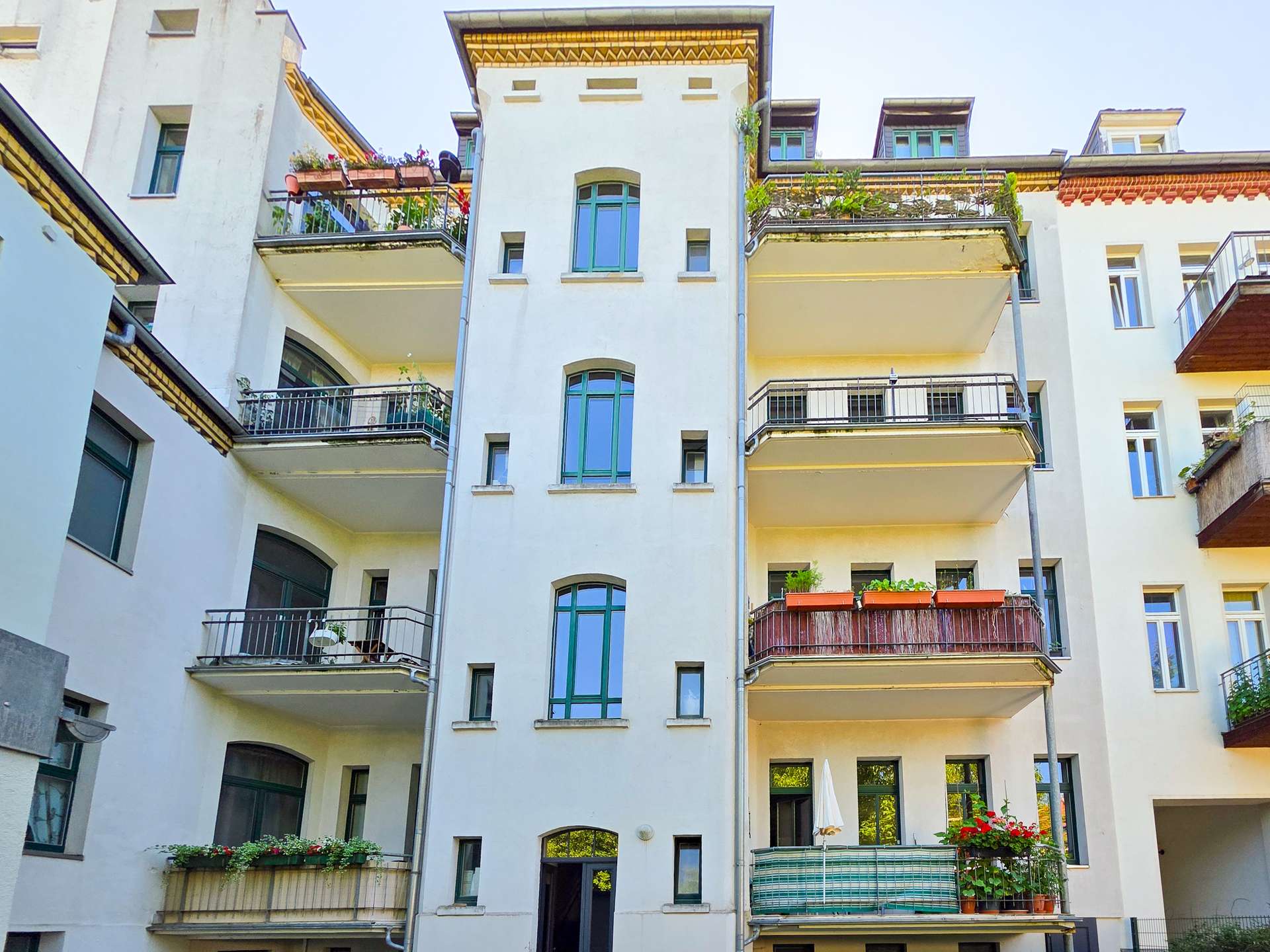 Ein weißes mehrstöckiges Wohnhaus mit Balkonen, die teilweise mit Pflanzen und Blumen geschmückt sind. Die Fassade ist mit großen Fenstern versehen, und auf beiden Seiten befinden sich Nebengebäude.