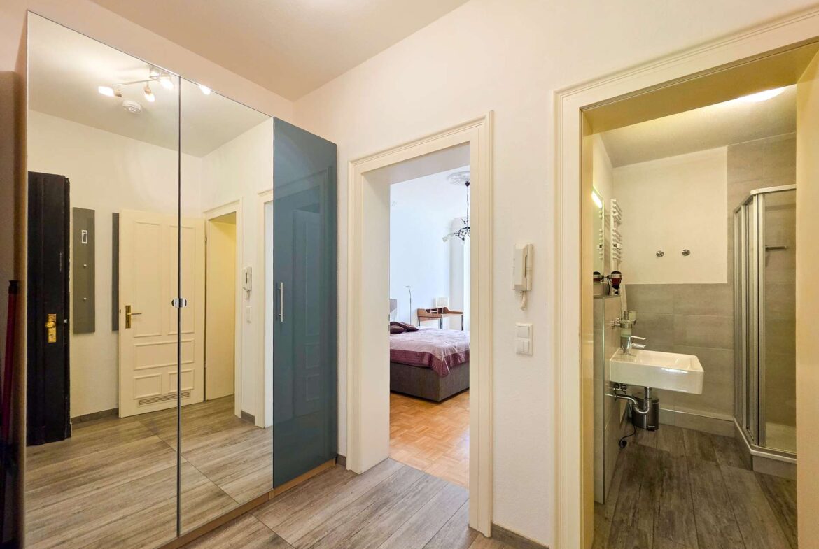 Ein Flur mit einem Spiegelschrank auf der linken Seite, der zu einem Schlafzimmer mit einem Bett und einem separaten Badezimmer mit Dusche, Waschbecken und Handtuchheizkörper auf der rechten Seite führt. Der Raum ist modern mit hellen Wänden und Holzfußböden.