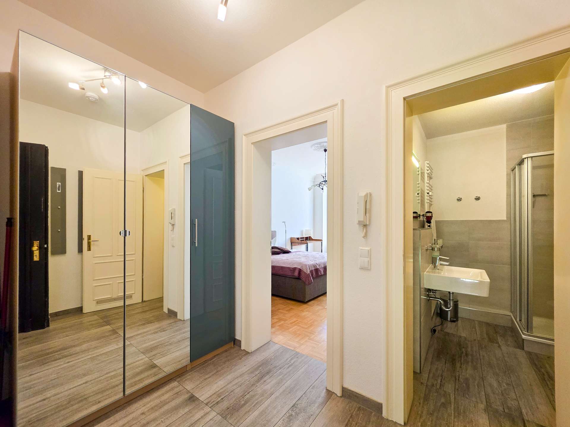 Ein Flur mit einem Spiegelschrank auf der linken Seite, der zu einem Schlafzimmer mit einem Bett und einem separaten Badezimmer mit Dusche, Waschbecken und Handtuchheizkörper auf der rechten Seite führt. Der Raum ist modern mit hellen Wänden und Holzfußböden.