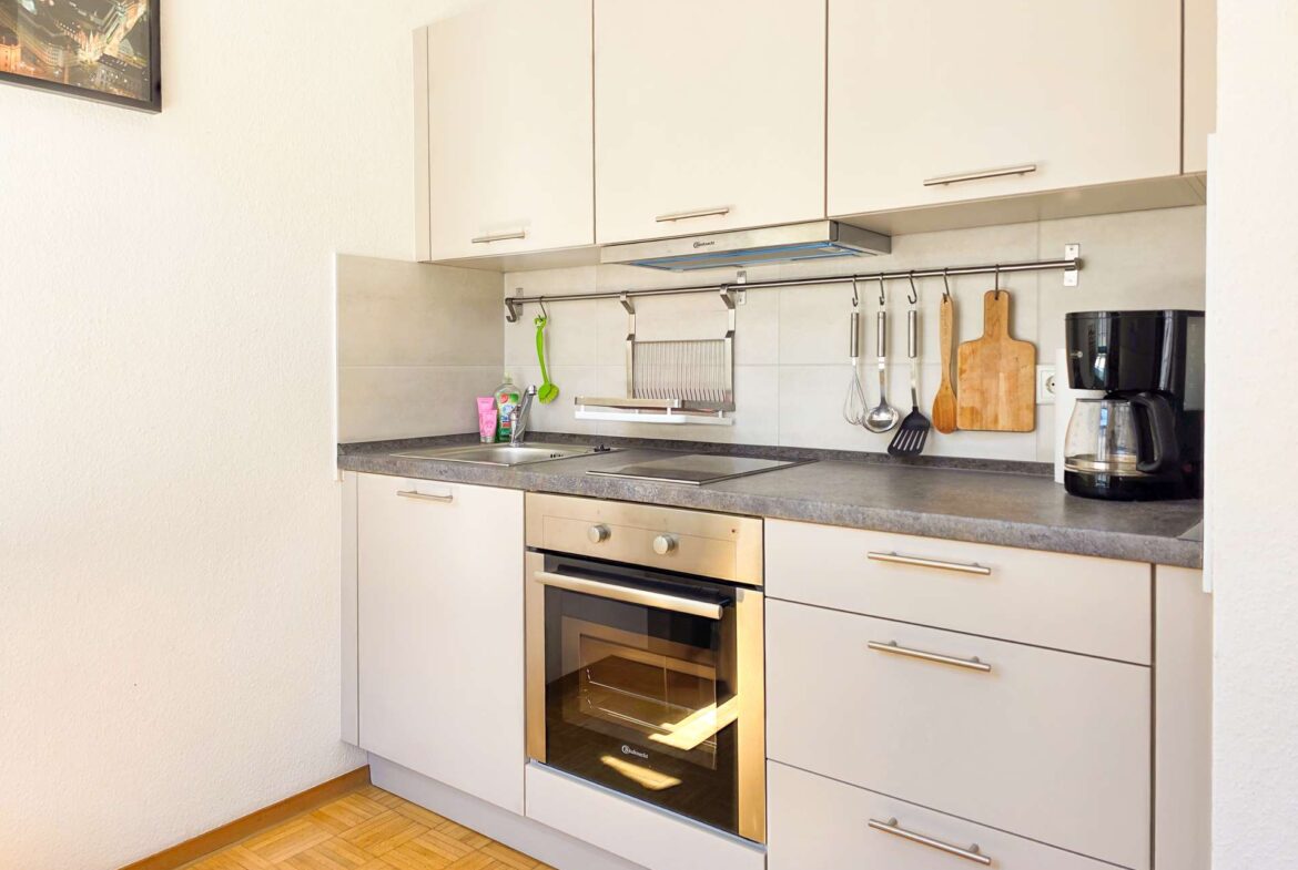 Moderne Küche mit weißen Schränken, einem Backofen, einer Spüle, einer Kaffeemaschine und Geschirr, das an einer Stange über der Theke hängt. Schneidebrett und Geschirrständer sind ebenfalls sichtbar. Der Raum ist sauber und gut organisiert.