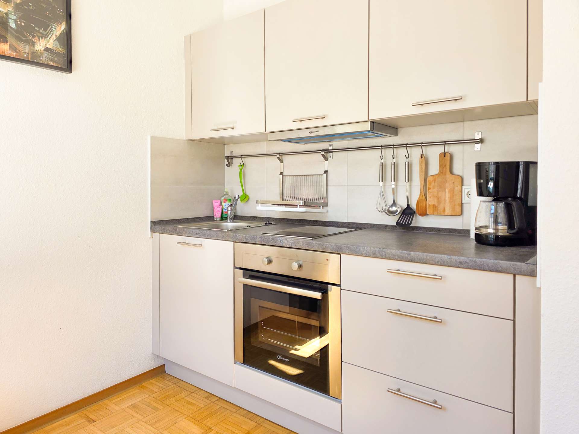 Moderne Küche mit weißen Schränken, einem Backofen, einer Spüle, einer Kaffeemaschine und Geschirr, das an einer Stange über der Theke hängt. Schneidebrett und Geschirrständer sind ebenfalls sichtbar. Der Raum ist sauber und gut organisiert.