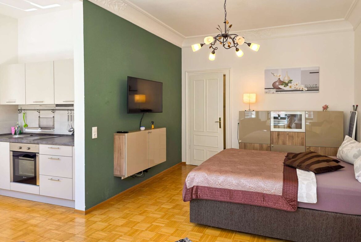 Ein modernes Studio-Apartment mit einem Bett, einem an der Wand montierten Fernseher und einer Küchenzeile. Das Zimmer verfügt über eine grüne Akzentwand, Parkettboden, eine Hängelampe und eine moderne Einrichtung.
