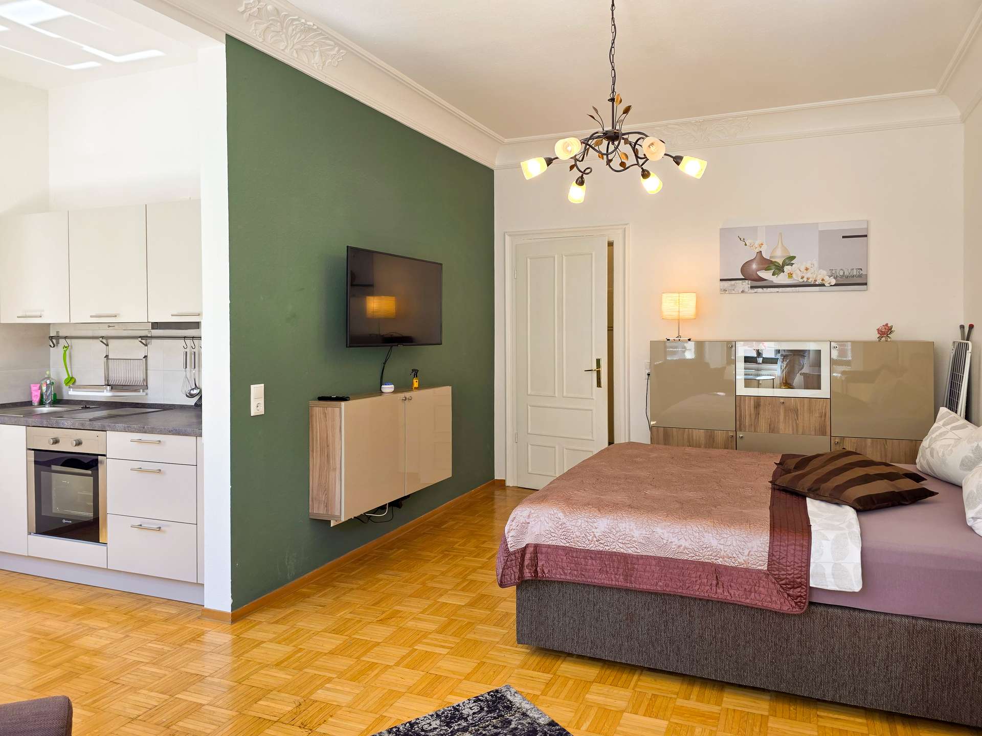 Ein modernes Studio-Apartment mit einem Bett, einem an der Wand montierten Fernseher und einer Küchenzeile. Das Zimmer verfügt über eine grüne Akzentwand, Parkettboden, eine Hängelampe und eine moderne Einrichtung.