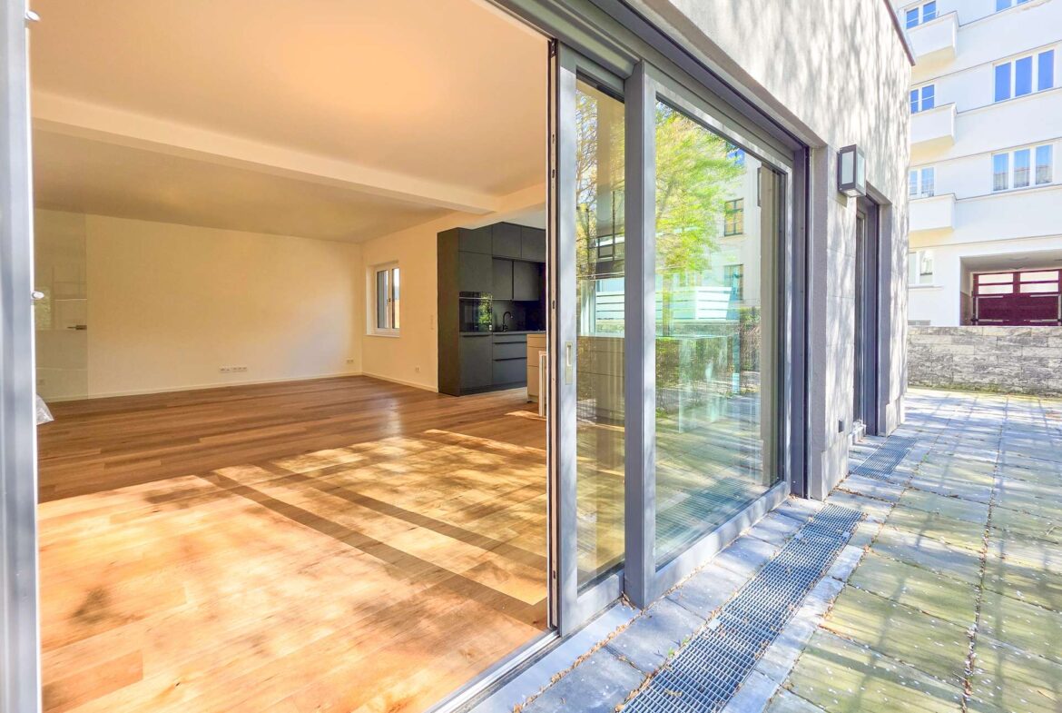 Open-plan living area with wooden floors, white walls, and a modern dark kitchen, opening to an outdoor patio through large glass sliding doors.