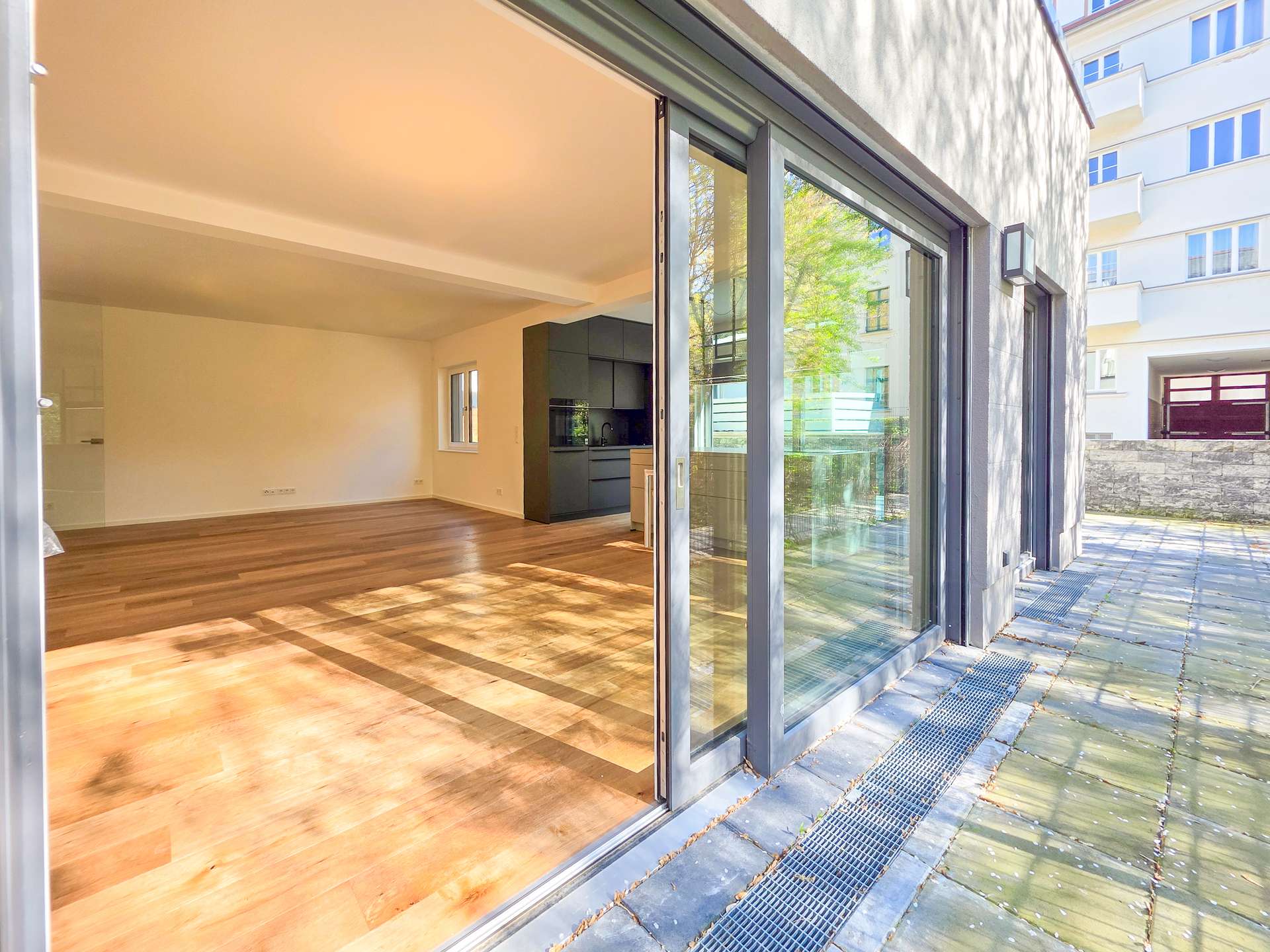 Open-plan living area with wooden floors, white walls, and a modern dark kitchen, opening to an outdoor patio through large glass sliding doors.