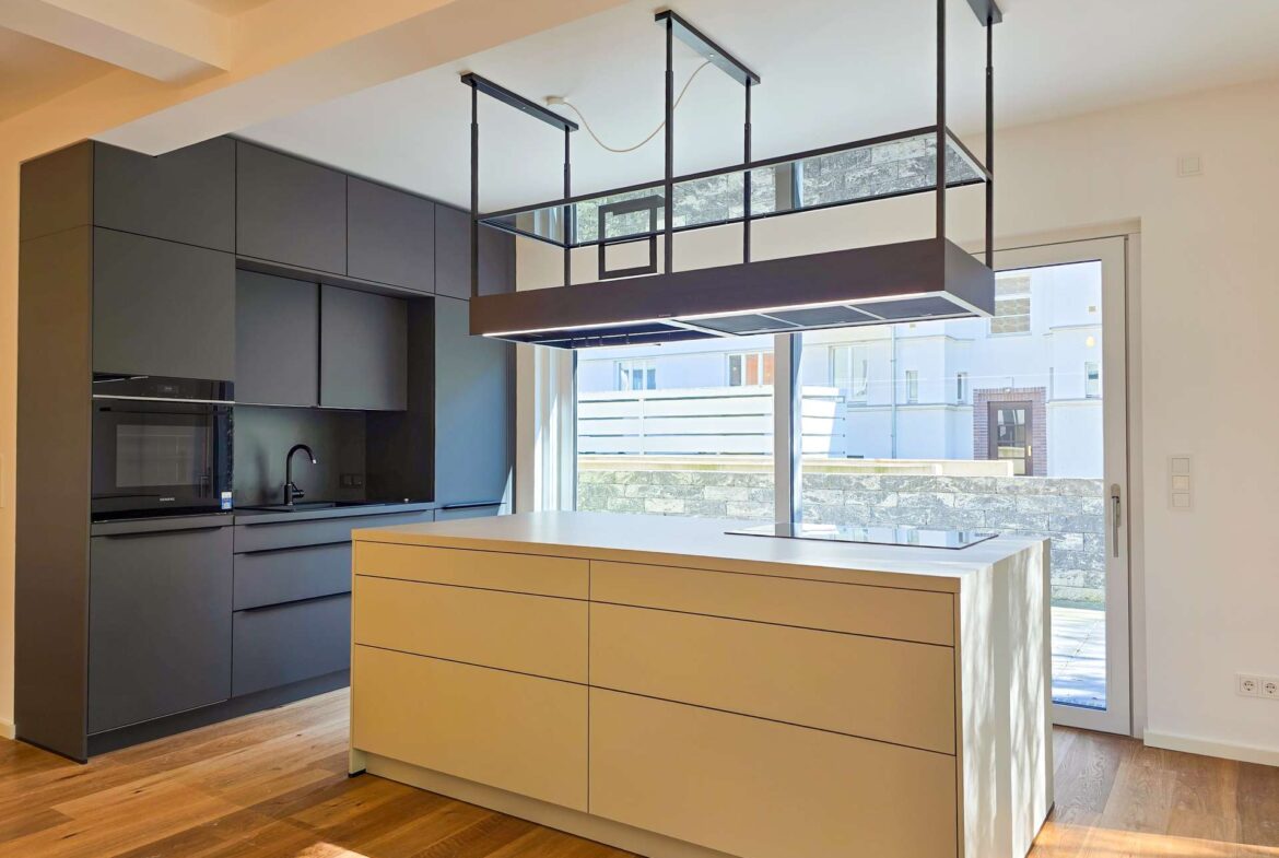 Contemporary kitchen with a white island, beige drawers, dark gray wall cabinets, built-in oven, and an overhead rectangular metal range hood over the island; large glass door lets in daylight.