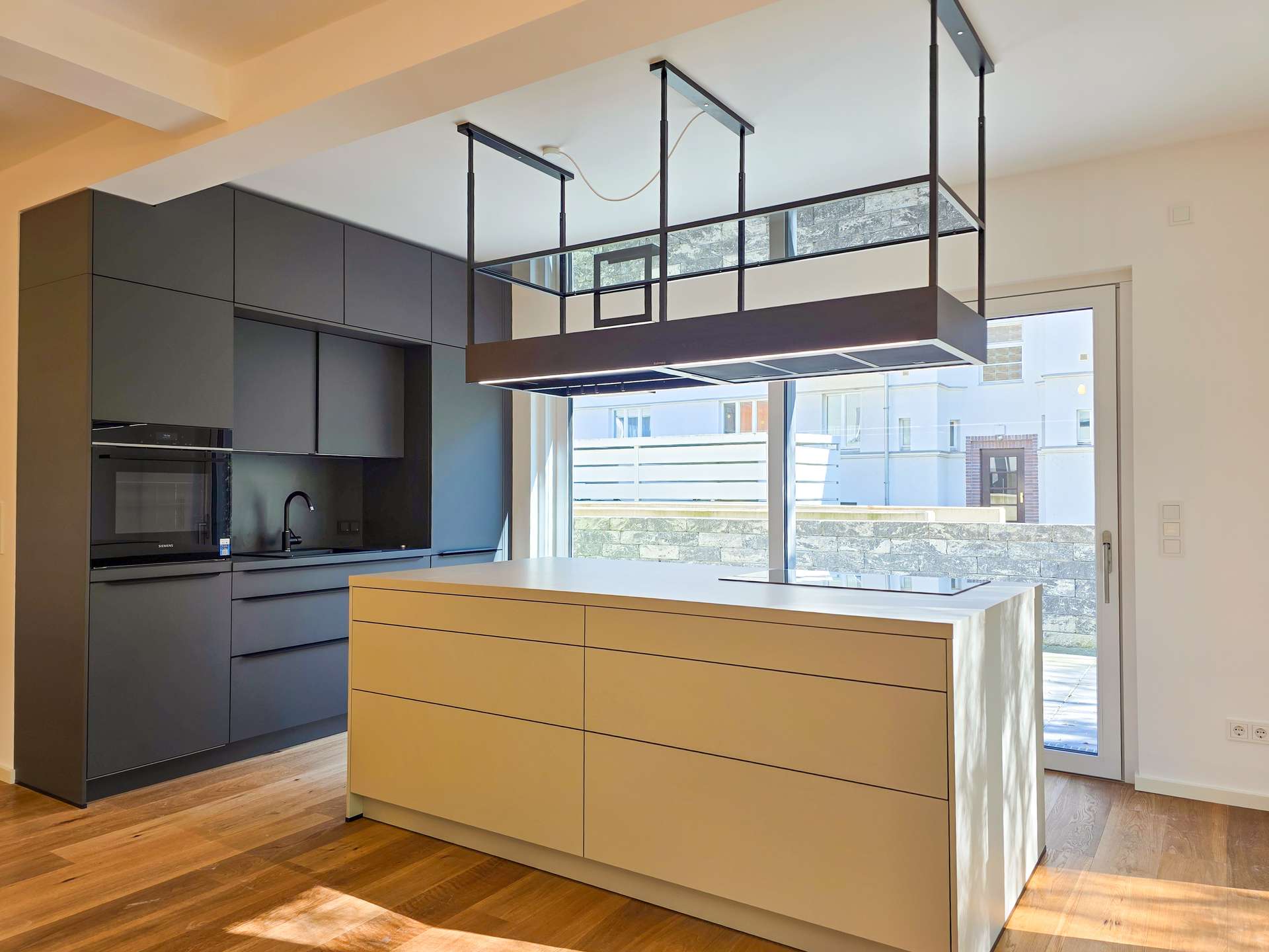 Contemporary kitchen with a white island, beige drawers, dark gray wall cabinets, built-in oven, and an overhead rectangular metal range hood over the island; large glass door lets in daylight.
