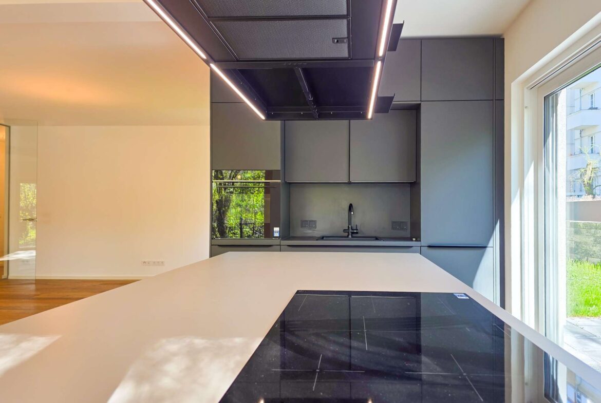 Contemporary kitchen with a white island, black induction cooktop, and dark gray cabinets under a ceiling-mounted range hood, with large windows bringing in daylight.
