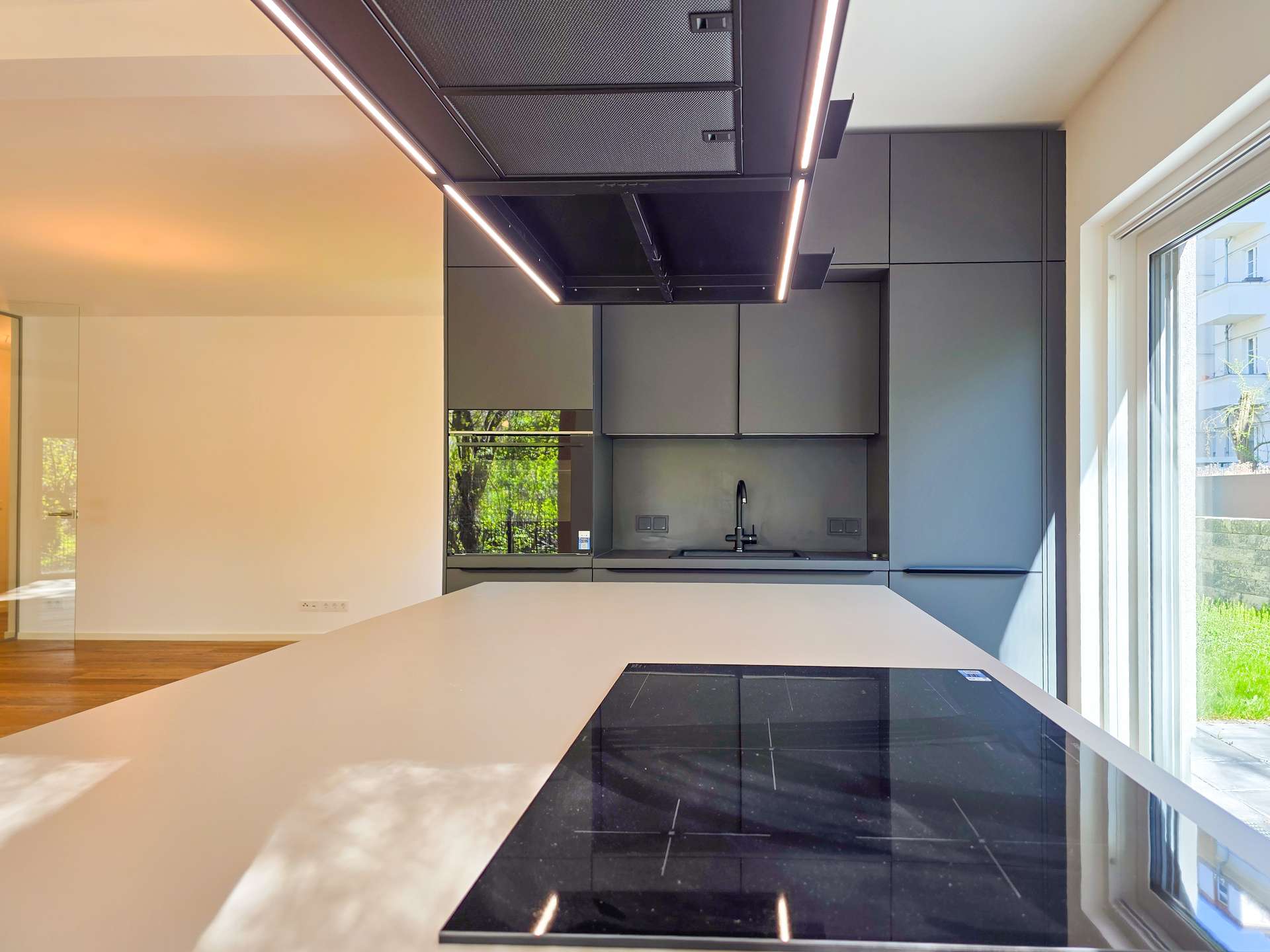 Contemporary kitchen with a white island, black induction cooktop, and dark gray cabinets under a ceiling-mounted range hood, with large windows bringing in daylight.