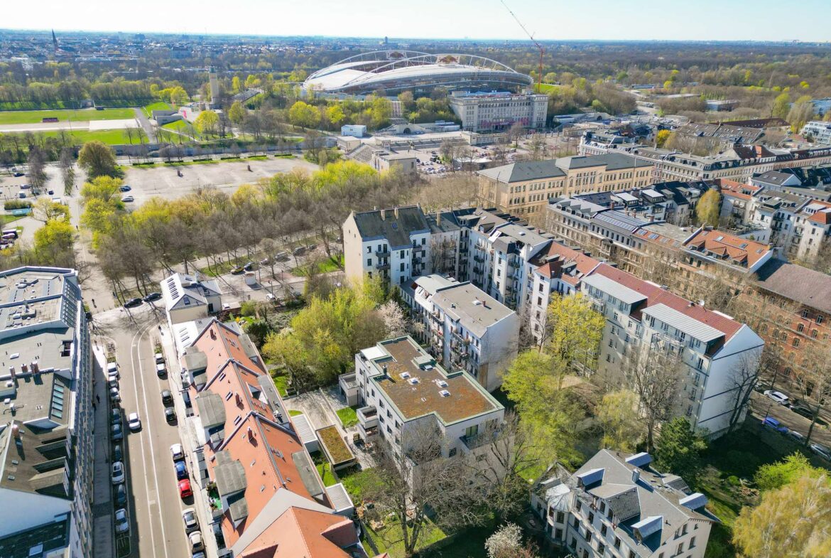 Aerial view of a city neighborhood with red-roofed buildings and a large futuristic stadium in the distance.