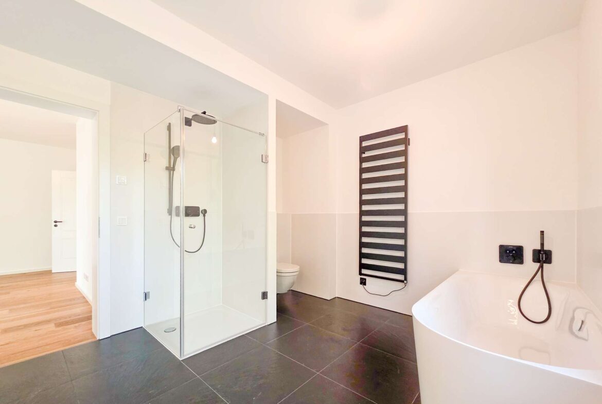 Modern bathroom with glass-enclosed shower, dark tile floor, and a white freestanding tub on the right.
