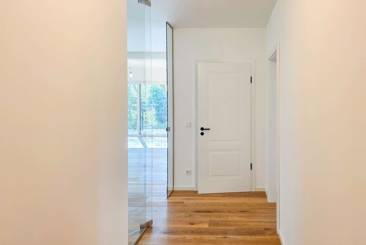 Bright hallway with a white paneled door at the end, glass left wall, and warm wooden flooring.