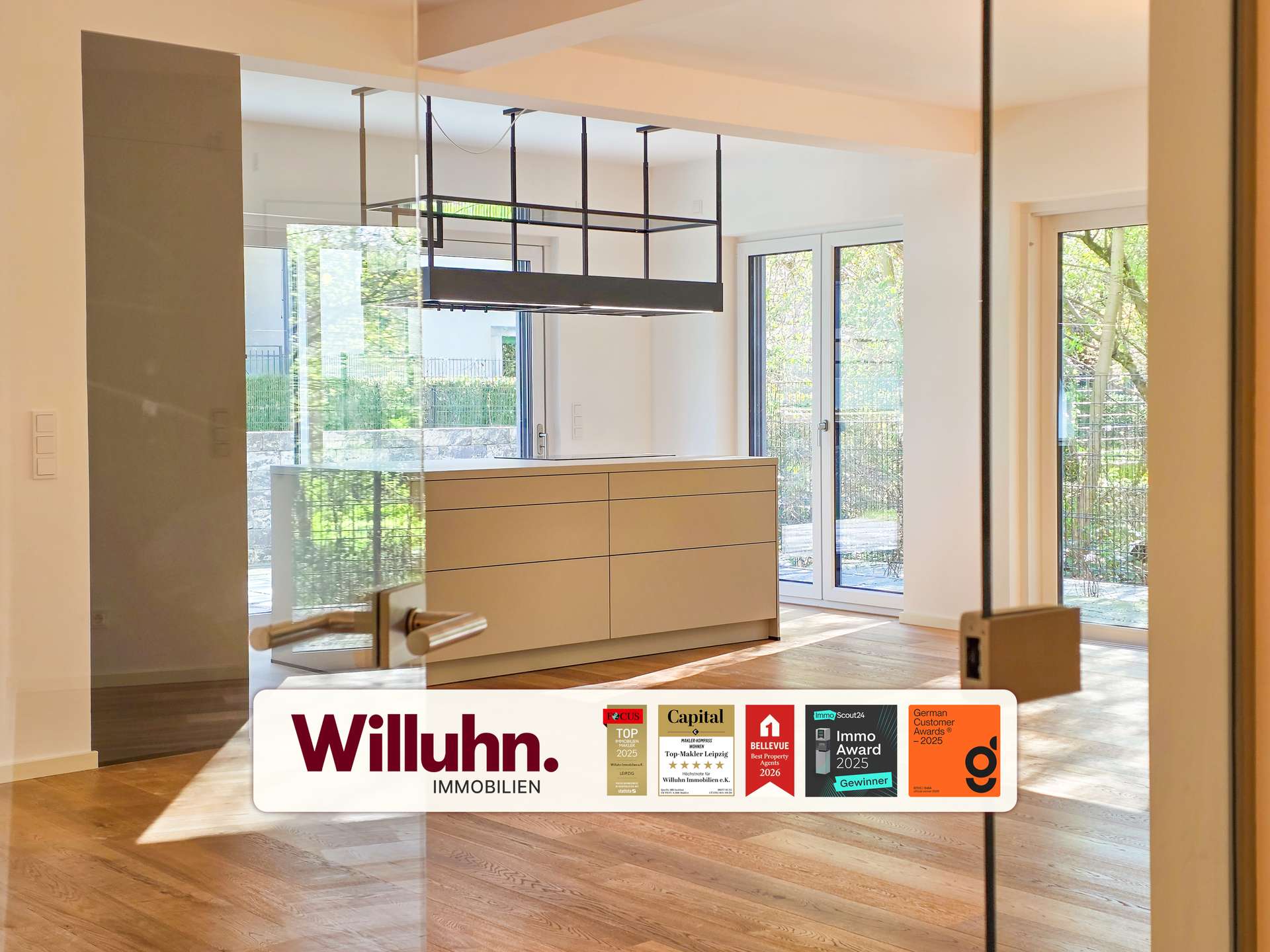 Modern showroom interior featuring beige cabinetry, glass walls, and large windows with natural light; Willuhn Immobilien branding in the foreground.