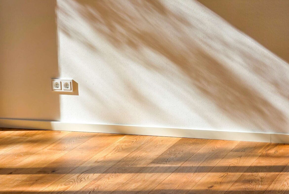 Interior wall with two electrical outlets near the left, sunlight casting diagonal shadows across the wall and wooden floor.