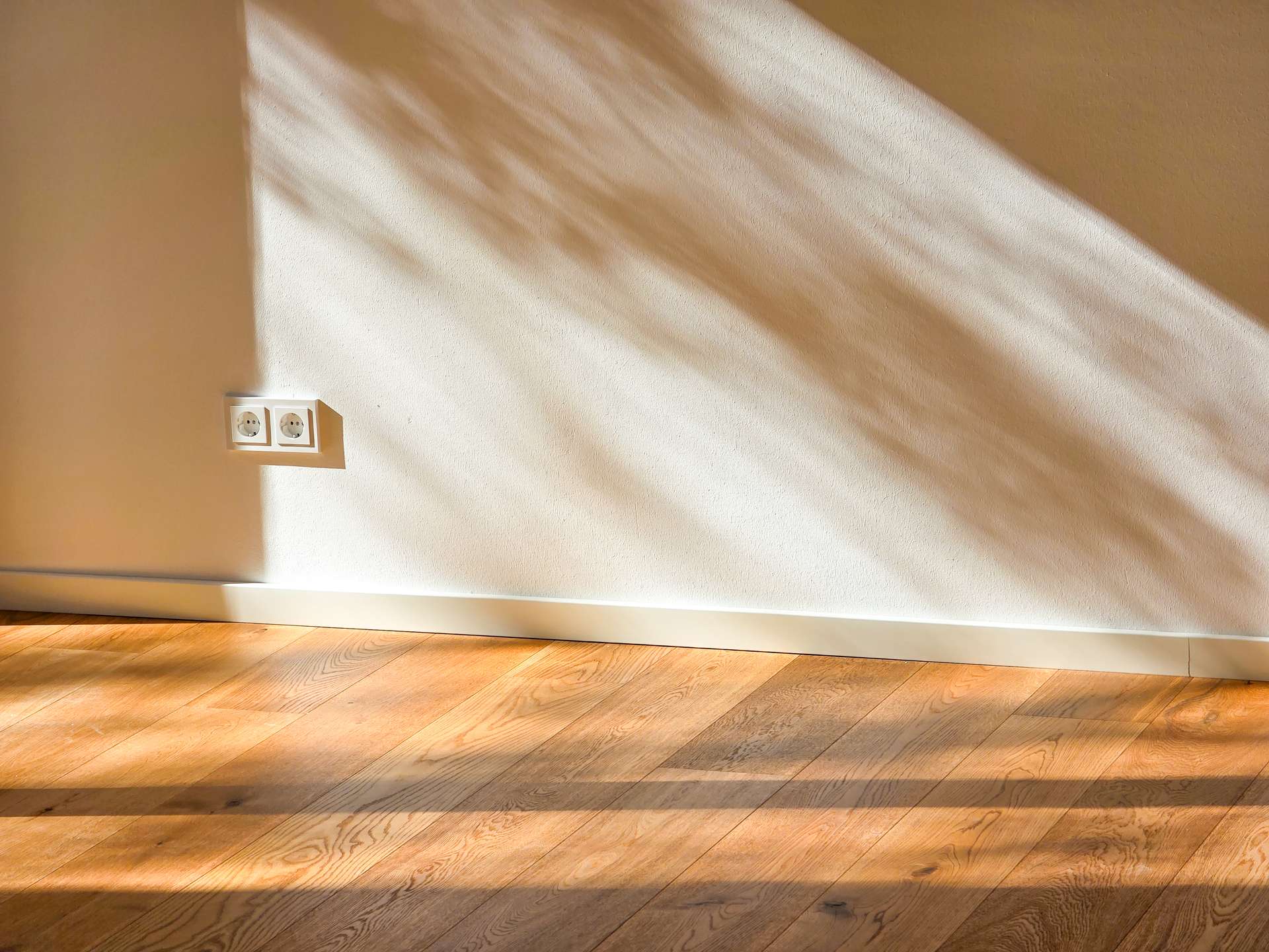 Interior wall with two electrical outlets near the left, sunlight casting diagonal shadows across the wall and wooden floor.