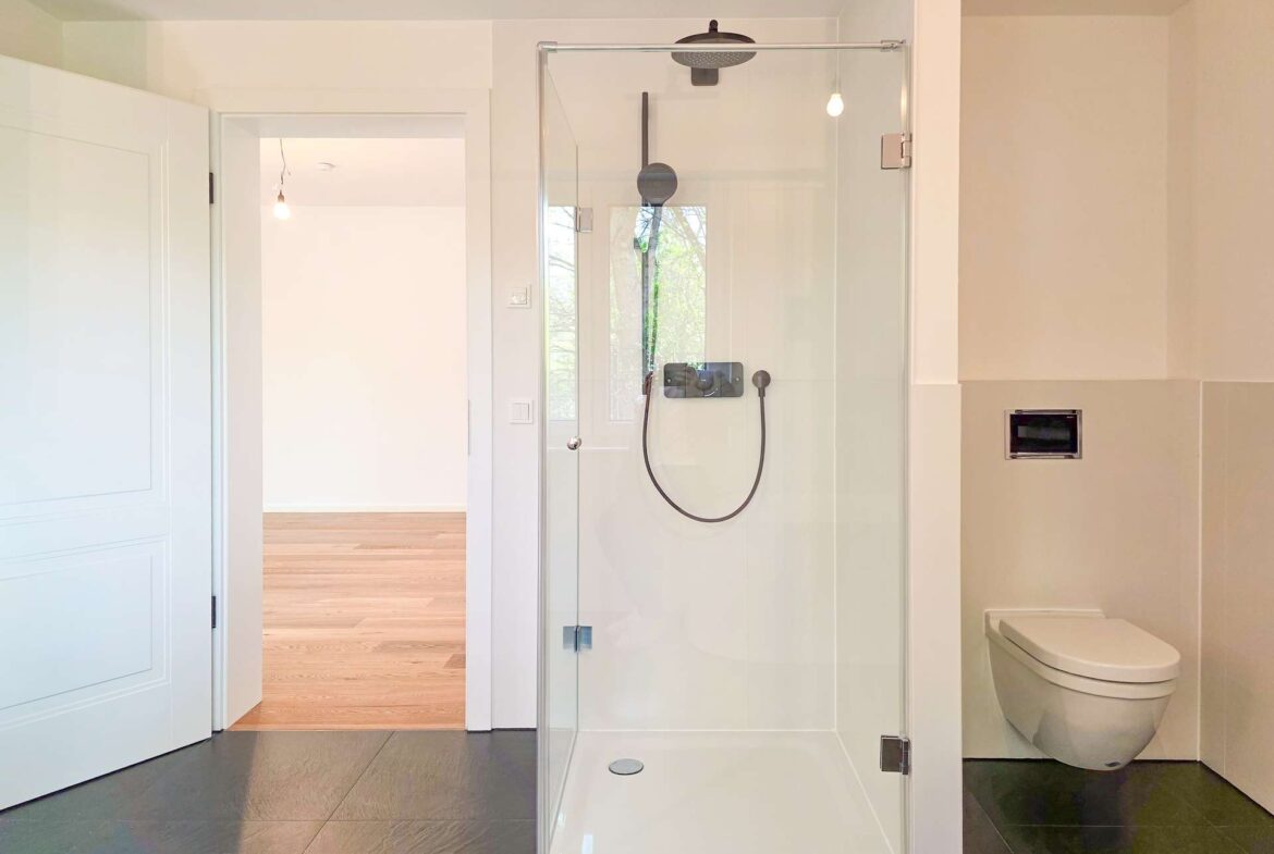 Modern bathroom with a frameless glass shower enclosure, black rainfall head, handheld wand, and wall controls; white toilet to the right and an open doorway to a bright hallway beyond.