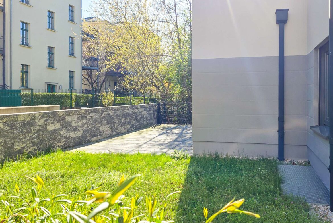 Courtyard scene with beige residential building on the left, stone wall and green hedge, sunlit trees in the center, and a pale building with a downspout on the right.