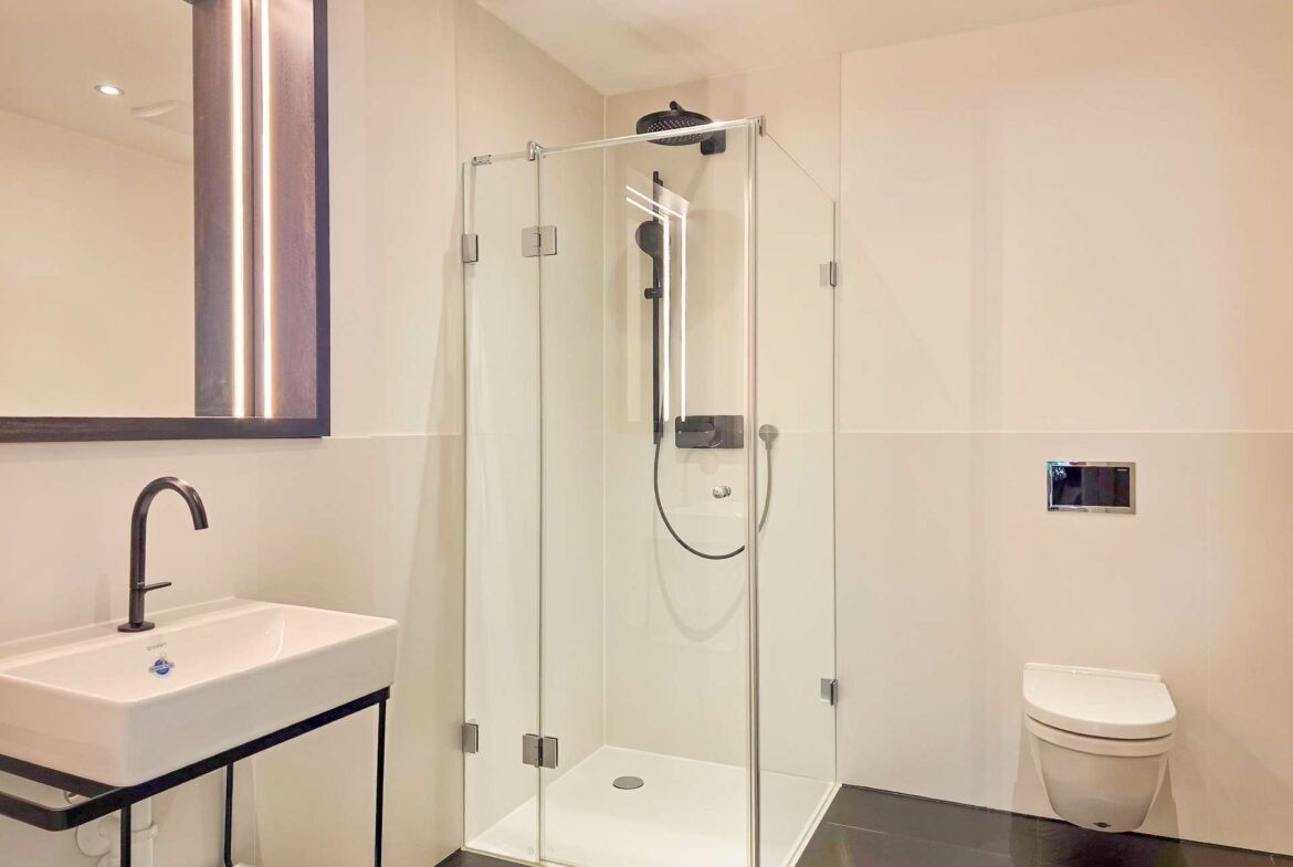 Modern bathroom with glass-walled shower, white sink with black faucet, and a wall-mounted toilet.