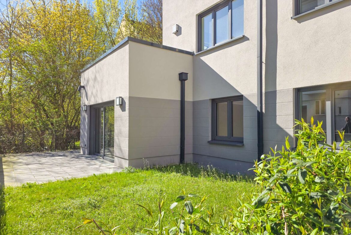 Modern beige apartment building with large windows, gray accents, and a paved patio area beside a green lawn.