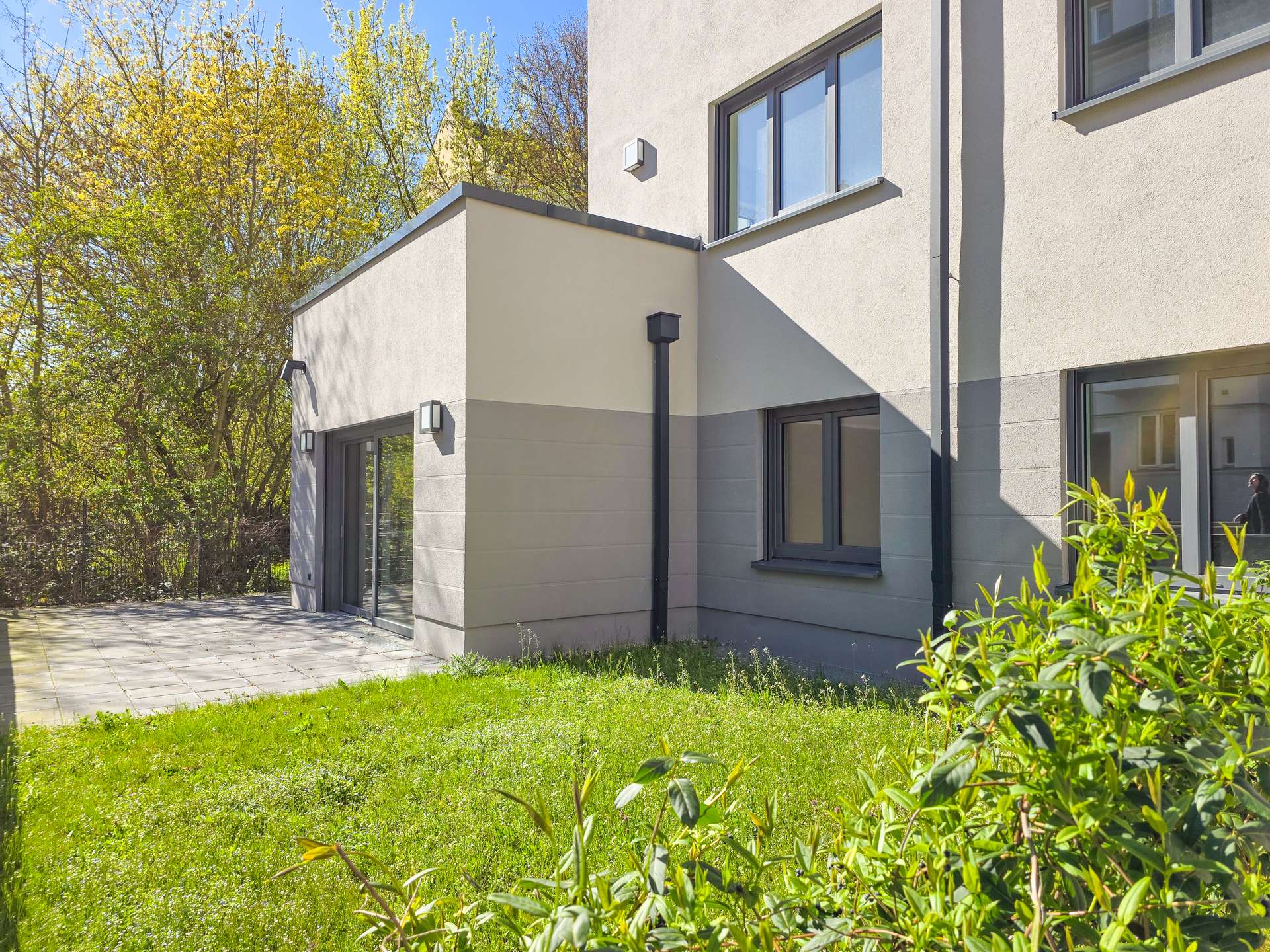Modern beige apartment building with large windows, gray accents, and a paved patio area beside a green lawn.