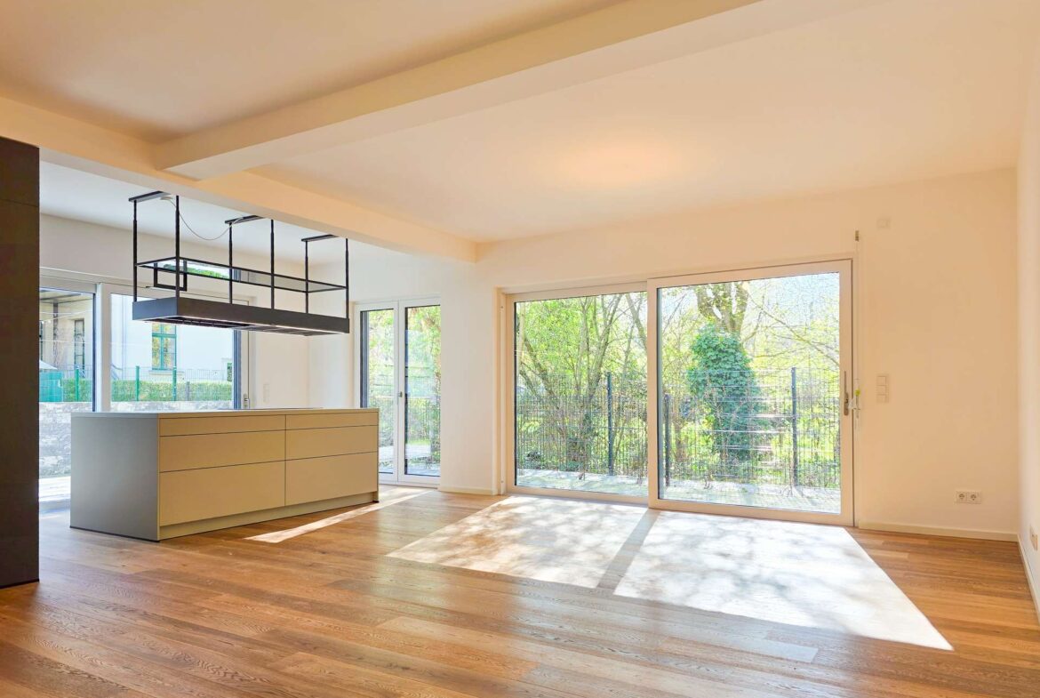 Open-plan kitchen and living area with warm wood flooring, white cabinetry, and large sliding glass doors overlooking a garden.