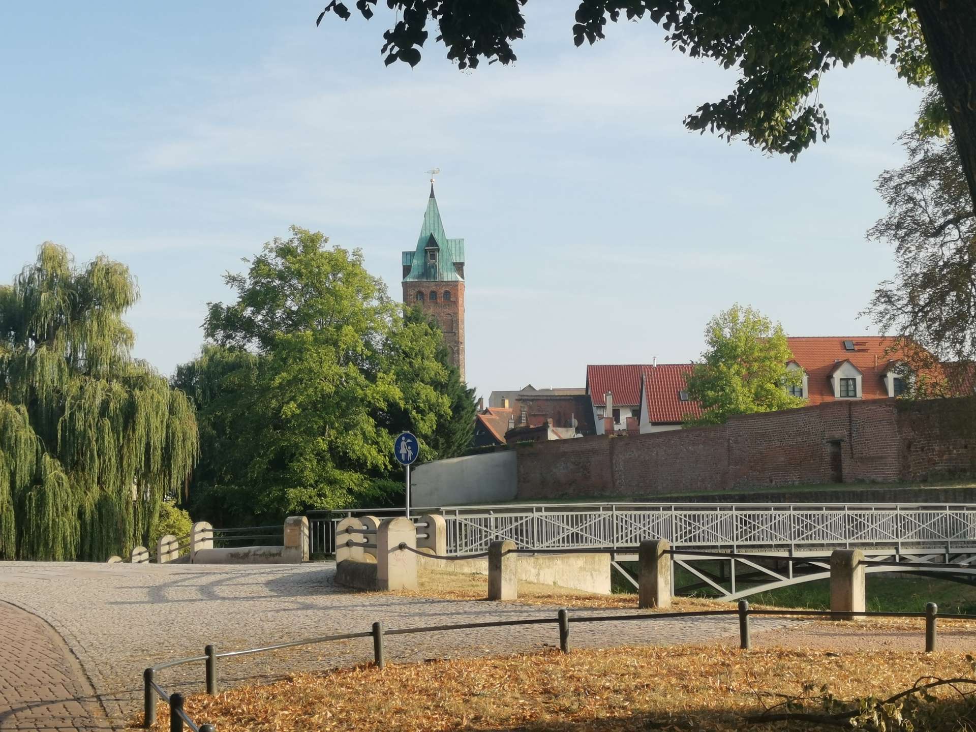 Brick church tower with a green copper roof peeking above trees along a cobblestone street.