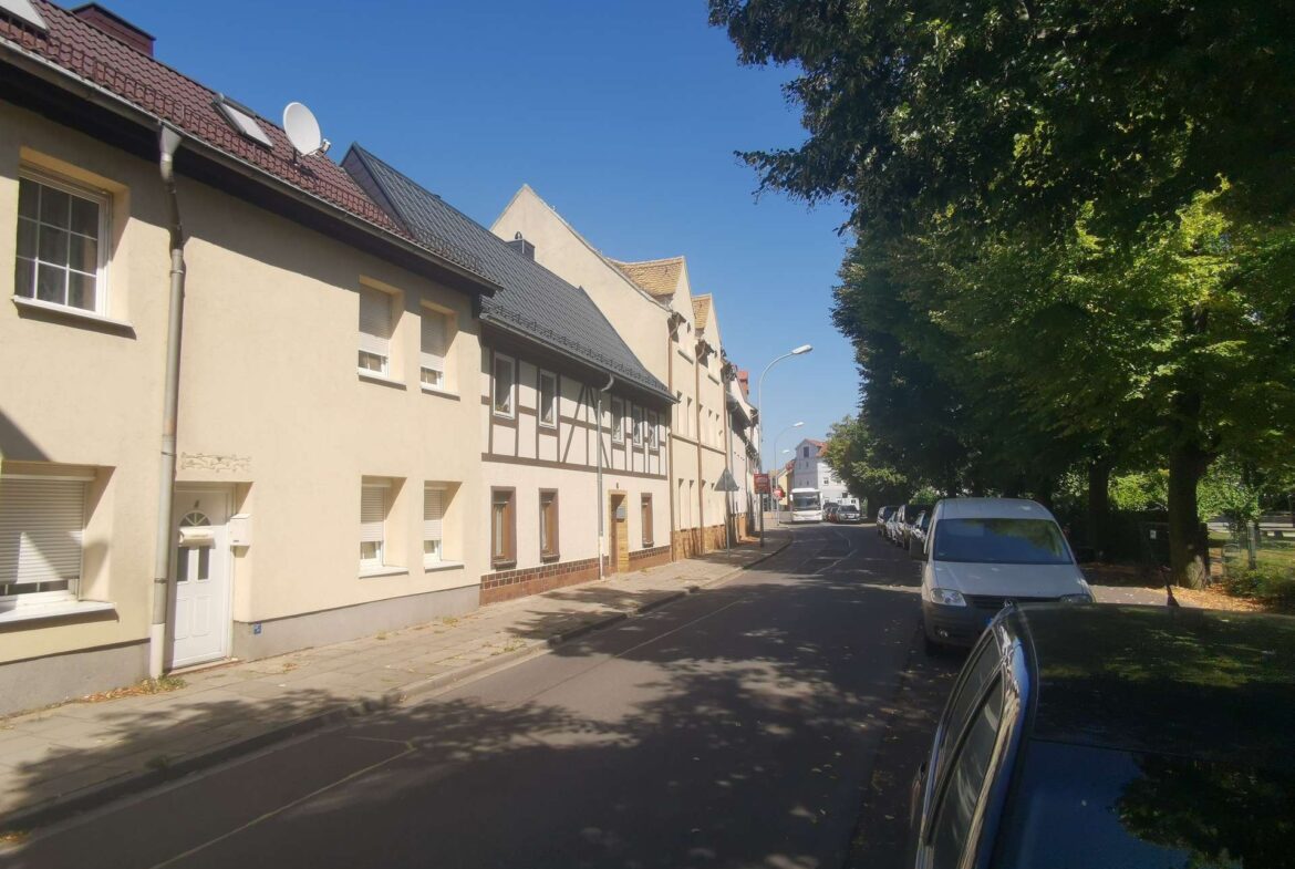 Residential street with beige row houses on the left, parked cars along the curb, and large green trees on the right under a clear blue sky.