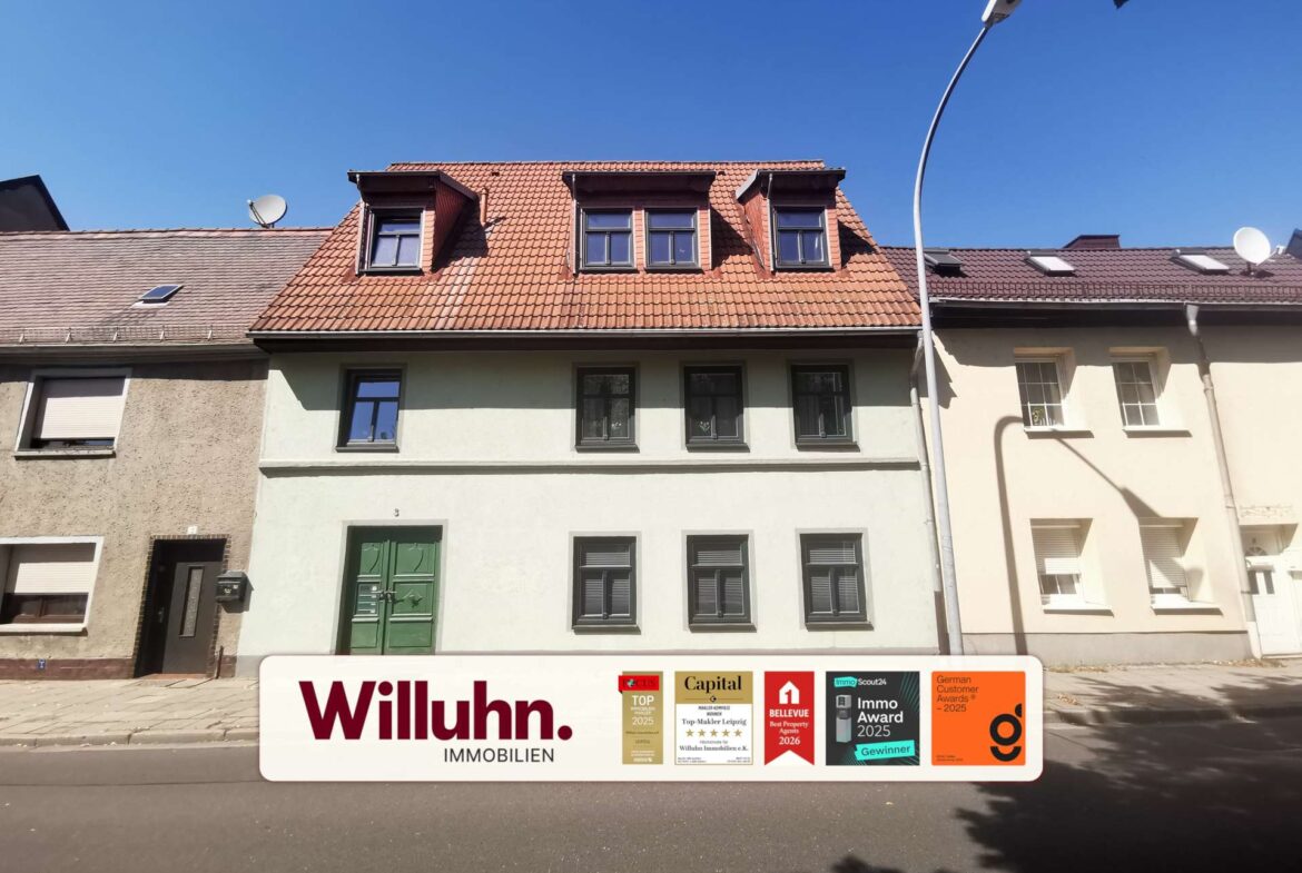 Facade of a rowhouse with red-tiled mansard roof and three dormer windows, under a clear blue sky; large Willuhn Immobilien sign with award badges visible in the foreground.