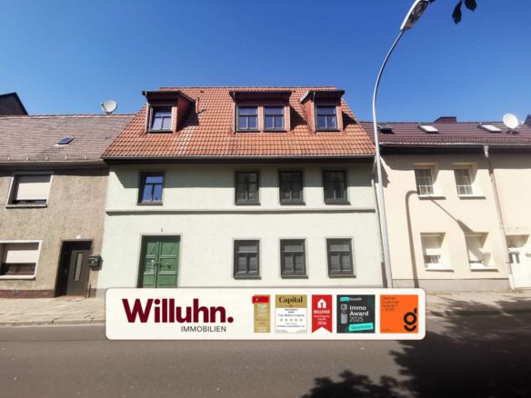 Facade of a rowhouse with red-tiled mansard roof and three dormer windows, under a clear blue sky; large Willuhn Immobilien sign with award badges visible in the foreground.