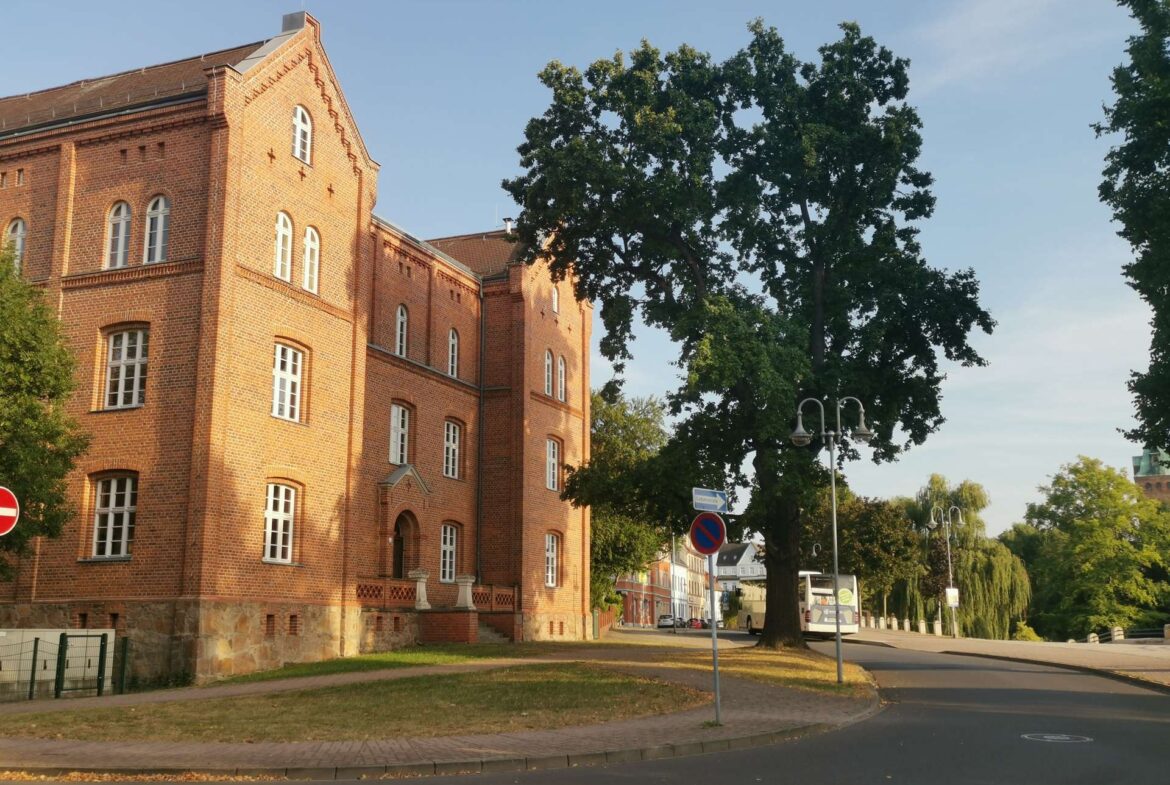 Red brick multi-story building with arched white-framed windows beside a large leafy tree on a sunny street corner.