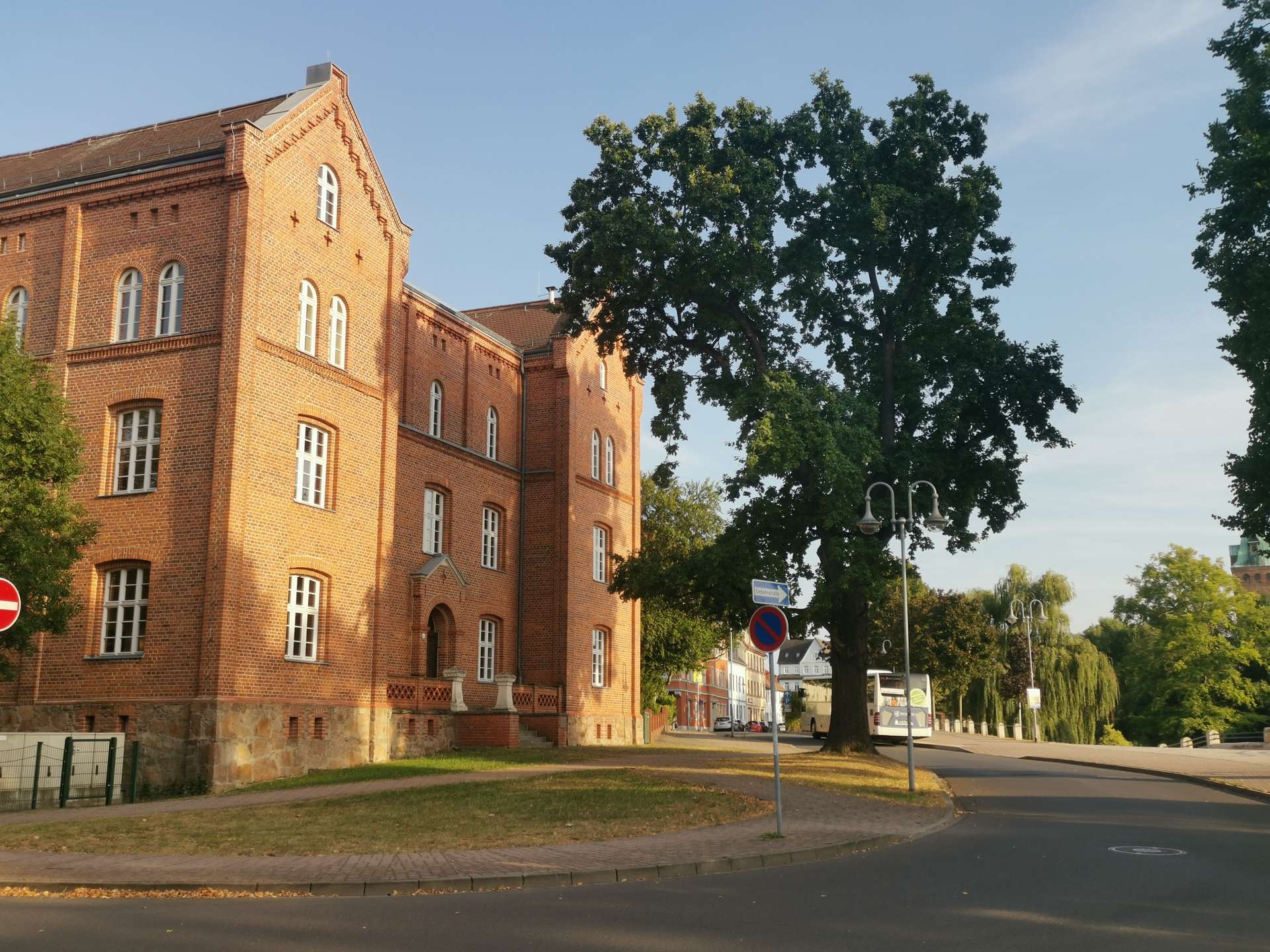 Red brick multi-story building with arched white-framed windows beside a large leafy tree on a sunny street corner.