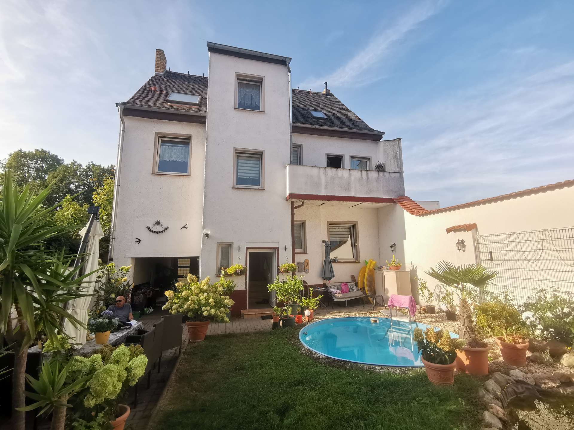 White multi-story house with a sloped roof and dormer windows, seen from a sunny backyard with a circular blue pool and potted plants.