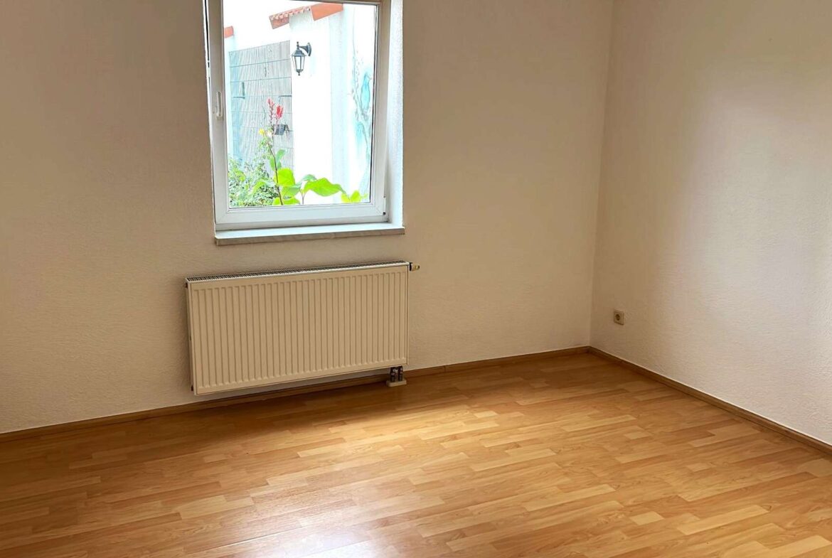 Empty small room with a window and a wall-mounted radiator beneath it, wooden floor, and a plant visible outside the window on the left side.
