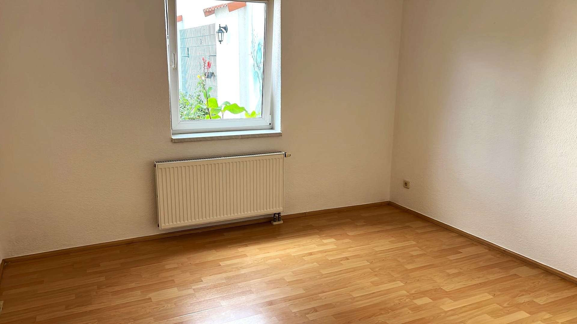 Empty small room with a window and a wall-mounted radiator beneath it, wooden floor, and a plant visible outside the window on the left side.