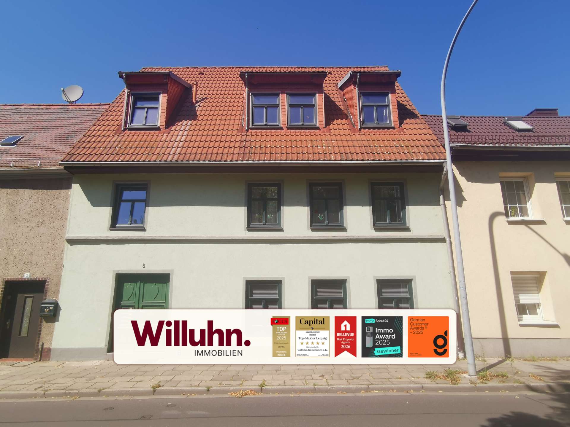 Front of a pale building with a red tiled roof and a large Willuhn Immobilien sign displaying award logos on the sidewalk in front of it.