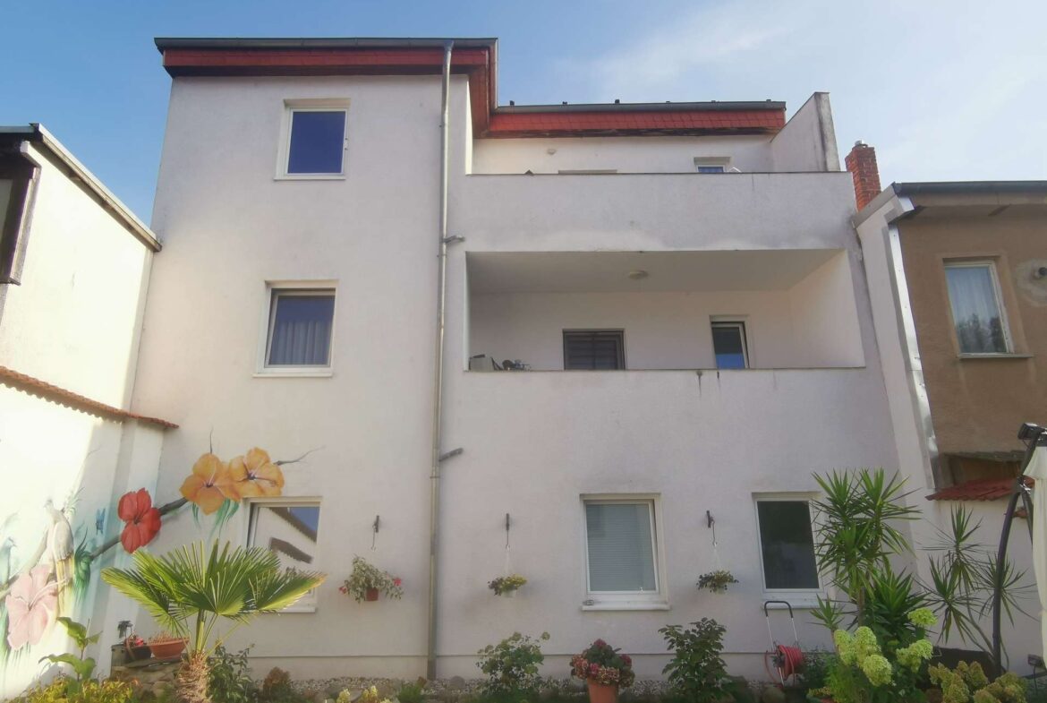 White multi-story apartment building with two balconies, a mural of flowers on the left wall, and a garden in front.