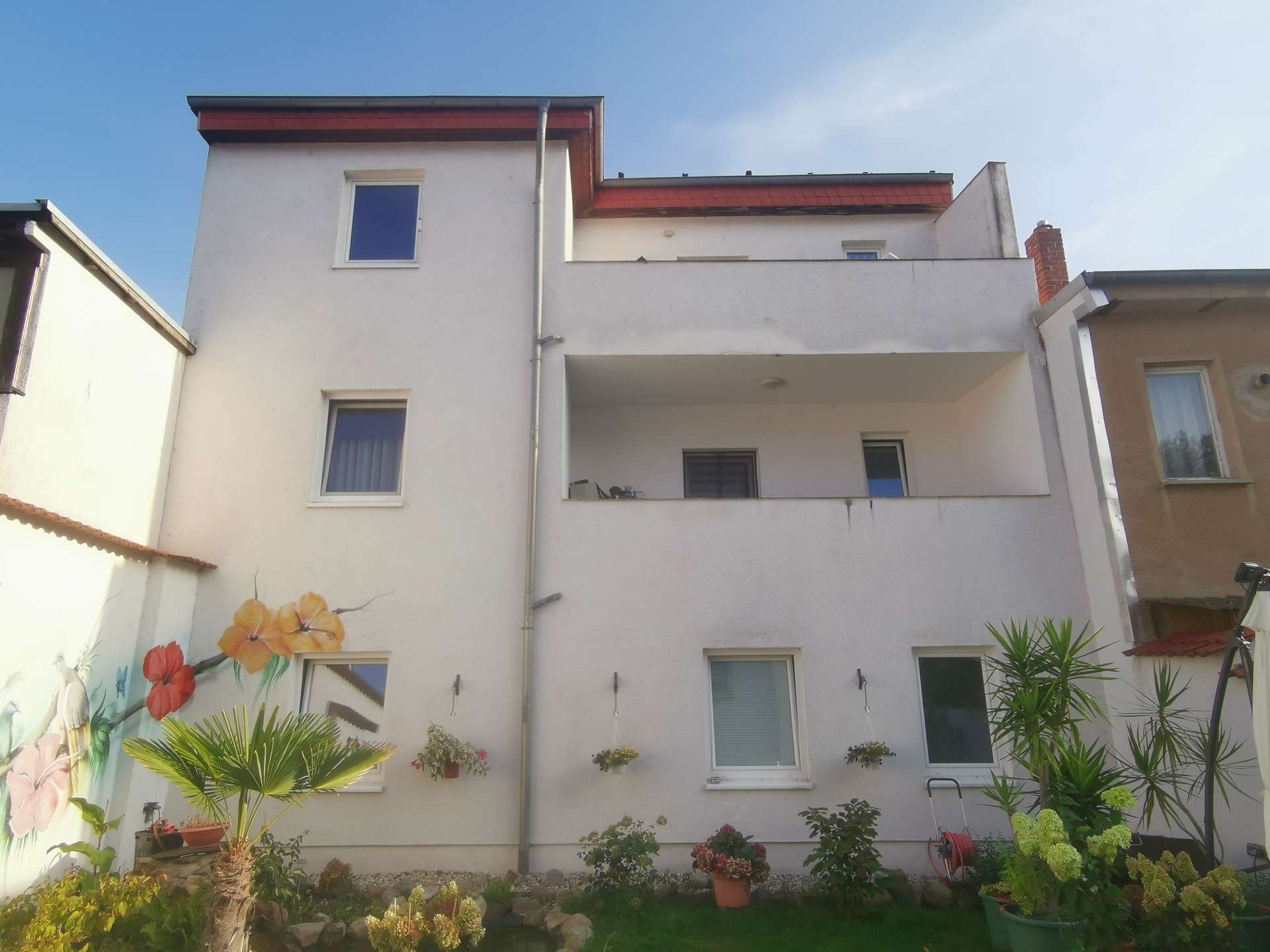 White multi-story apartment building with two balconies, a mural of flowers on the left wall, and a garden in front.