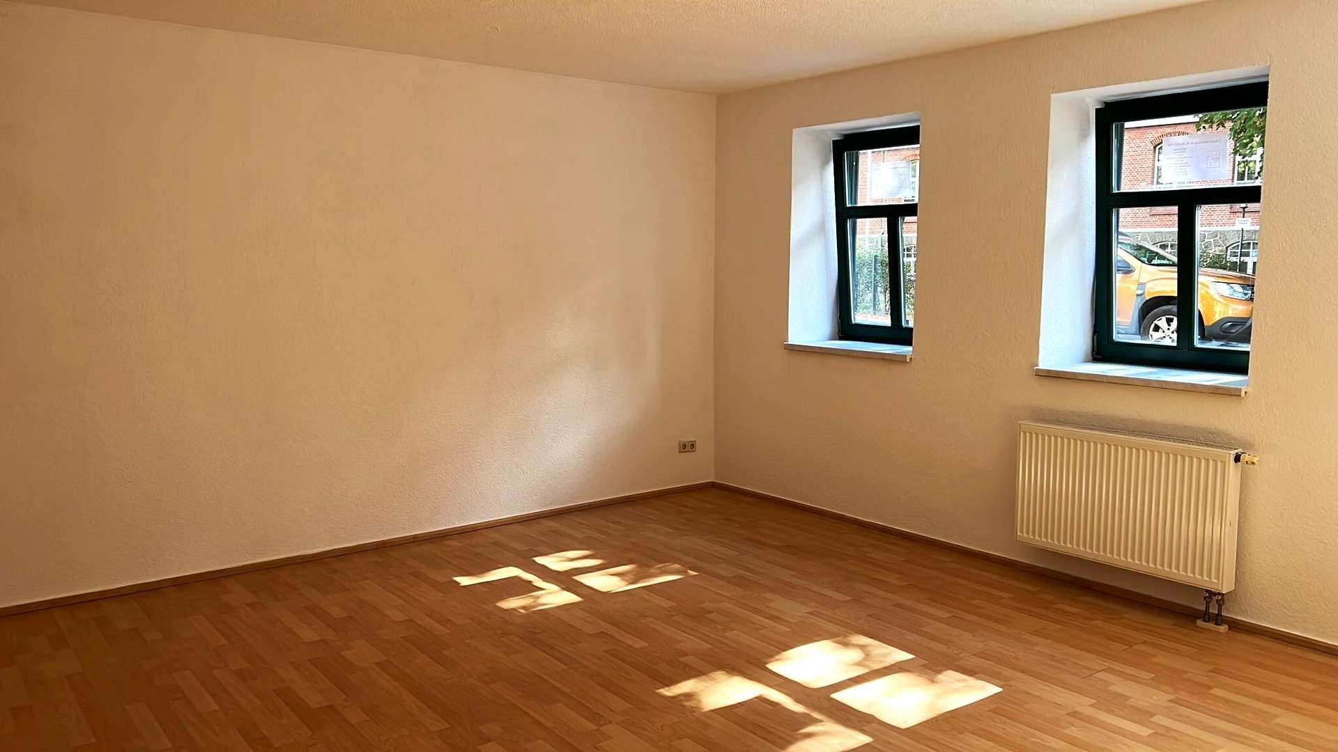 Empty room with light wood floor, white walls, and two dark-framed windows letting in daylight; radiator along the right wall.