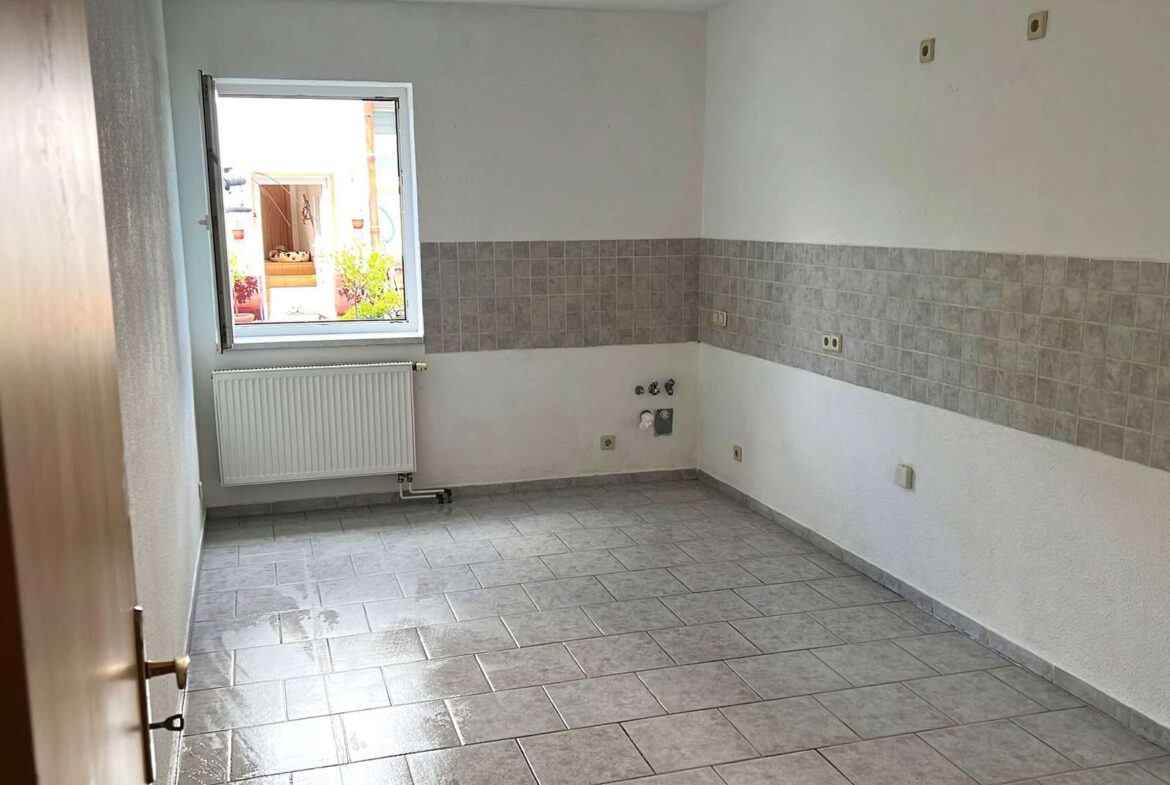 Empty kitchen with tiled floor and half-tiled backsplash, window open to outdoor courtyard, radiator under window, plumbing hookups on wall.